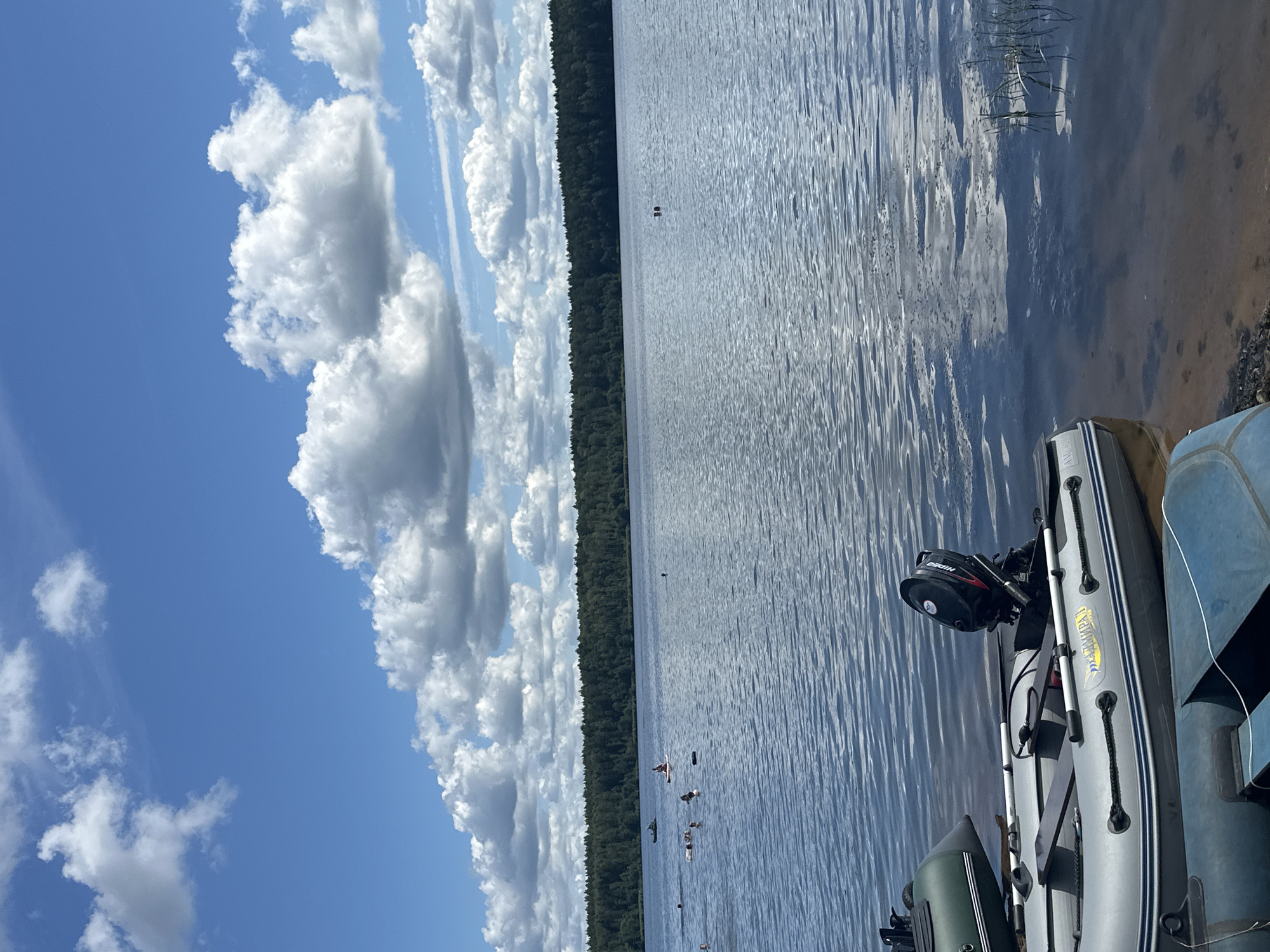 A group of people are swimming and paddleboarding in a calm lake on a sunny day. In the foreground, several inflatable boats are parked on the shore. The lake is surrounded by a dense forest. The sky is blue with scattered white clouds.