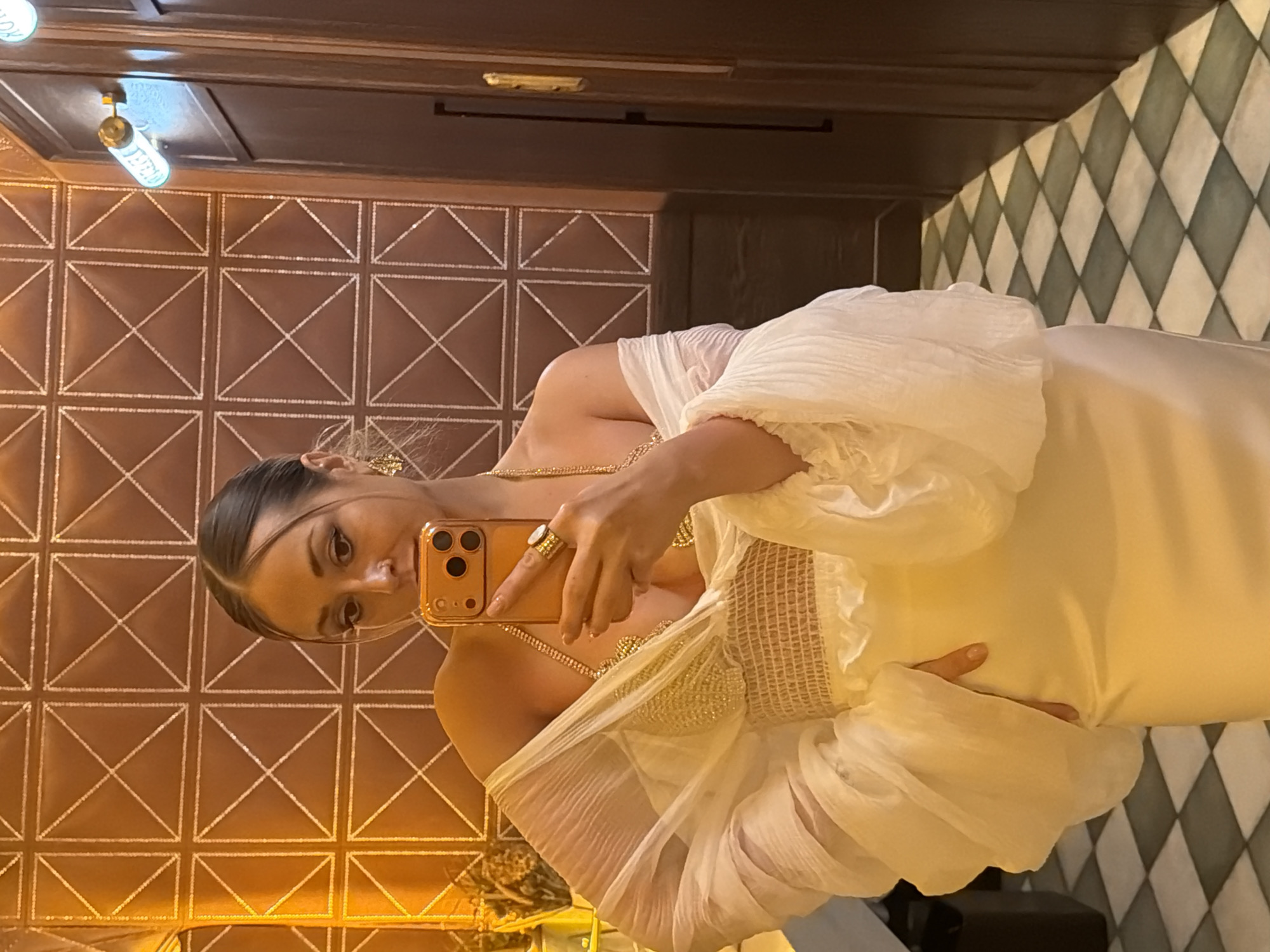 A woman captures a mirror selfie in an upscale interior setting, located in Krasnoyarsk, Russia. She has dark hair pulled back and wears visible makeup, including a nose ring. Her outfit consists of a gold, beaded bra-top, layered under a sheer, off-white, off-the-shoulder garment with voluminous, pleated sleeves and a smocked waist. The lower part of her dress or skirt is a smooth, light-colored fabric. She wears large gold earrings, multiple gold necklaces, and a ring with a white stone on her right hand, which holds an orange-cased smartphone with a triple camera lens. Her left hand rests on her hip. The background features a patterned wall upholstered in brown material with gold stitching forming a grid of squares, each containing an 'X' pattern, bathed in warm, golden light. To the right, a dark wooden door with a vertical handle is visible, along with a light fixture emitting a blue glow (possibly a neon sign) and a smaller golden fixture. The floor is covered in a checkerboard pattern of alternating light and dark green diamond-shaped tiles.