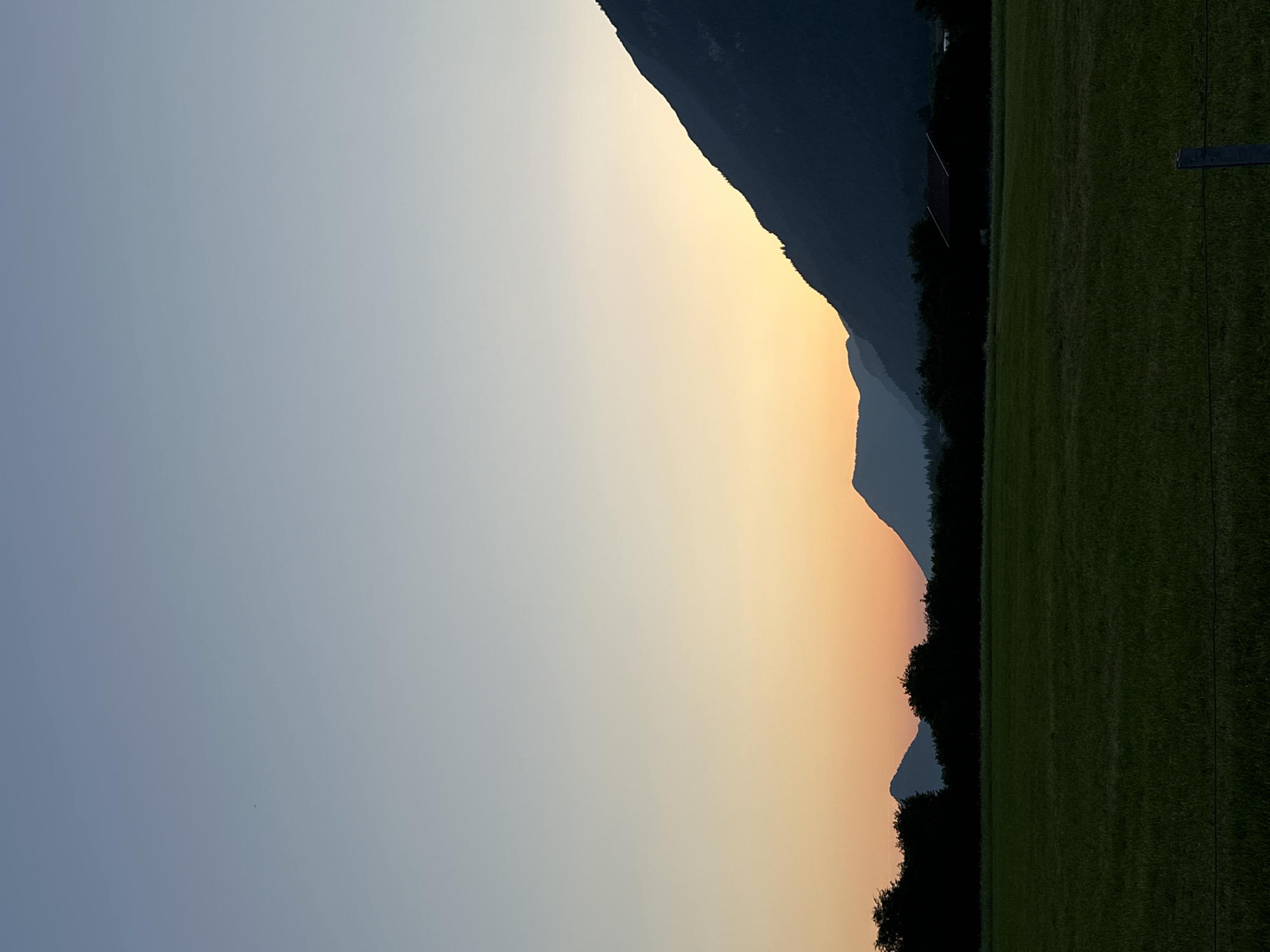 This image depicts a serene sunset scene with mountains silhouetted against a gradient sky of pale blue and warm orange. In the foreground, a dark green field stretches towards the horizon, with a thin wire fence running across the bottom of the frame. Behind the field, a dense line of dark trees obscures the base of the mountains. A dark, angular structure, possibly a barn or shed, is visible nestled within the trees on the right side of the image. The overall impression is one of tranquility and natural beauty, suggesting a rural setting near Garmisch-Partenkirchen, Germany, known for its picturesque mountain landscapes.