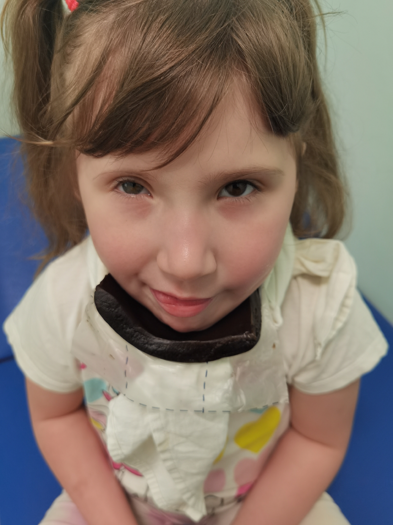 A young girl with brown hair in pigtails, wearing a white t-shirt and a bib with a dark brown lining, is sitting indoors on a blue surface. The girl is looking directly at the camera with a slight smile, and her tongue is slightly visible. The background is a light blue or green color. The location is identified as Perm, Russia based on other information.