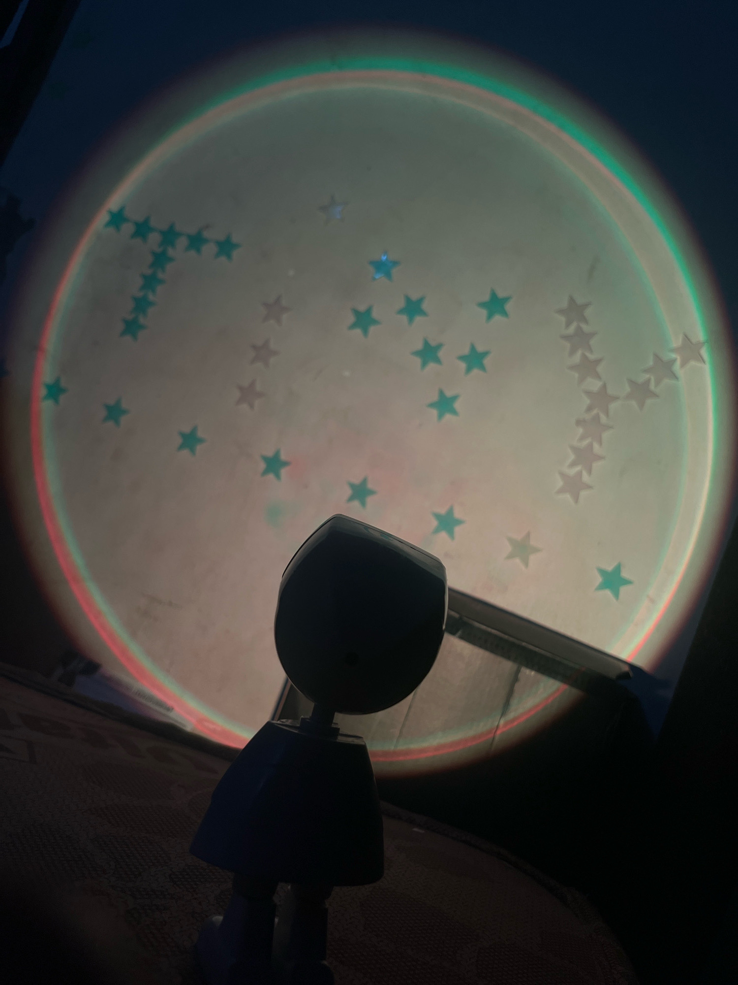 A small, dark, toy figure with a rounded head and a blue, flared skirt is positioned in the foreground, facing away from the camera and towards a large, luminous circle projected onto a surface. This projected circle displays a gradient of colors, beginning with red on the bottom left and transitioning through orange, yellow, green, and blue towards the top. Scattered across the projected circle are numerous small, star-shaped figures. Some stars are a light blue or teal color, while others are a pale, almost white, hue. The toy figure appears to be standing on a textured surface, possibly a rug or mat, with other dimly lit objects or surfaces visible in the background. The overall impression is that of a decorative light projection creating a starry, circular effect, with the toy figure as a solitary observer. This scene is not specifically identifiable as being in Lokoja, Nigeria, or any other particular city.