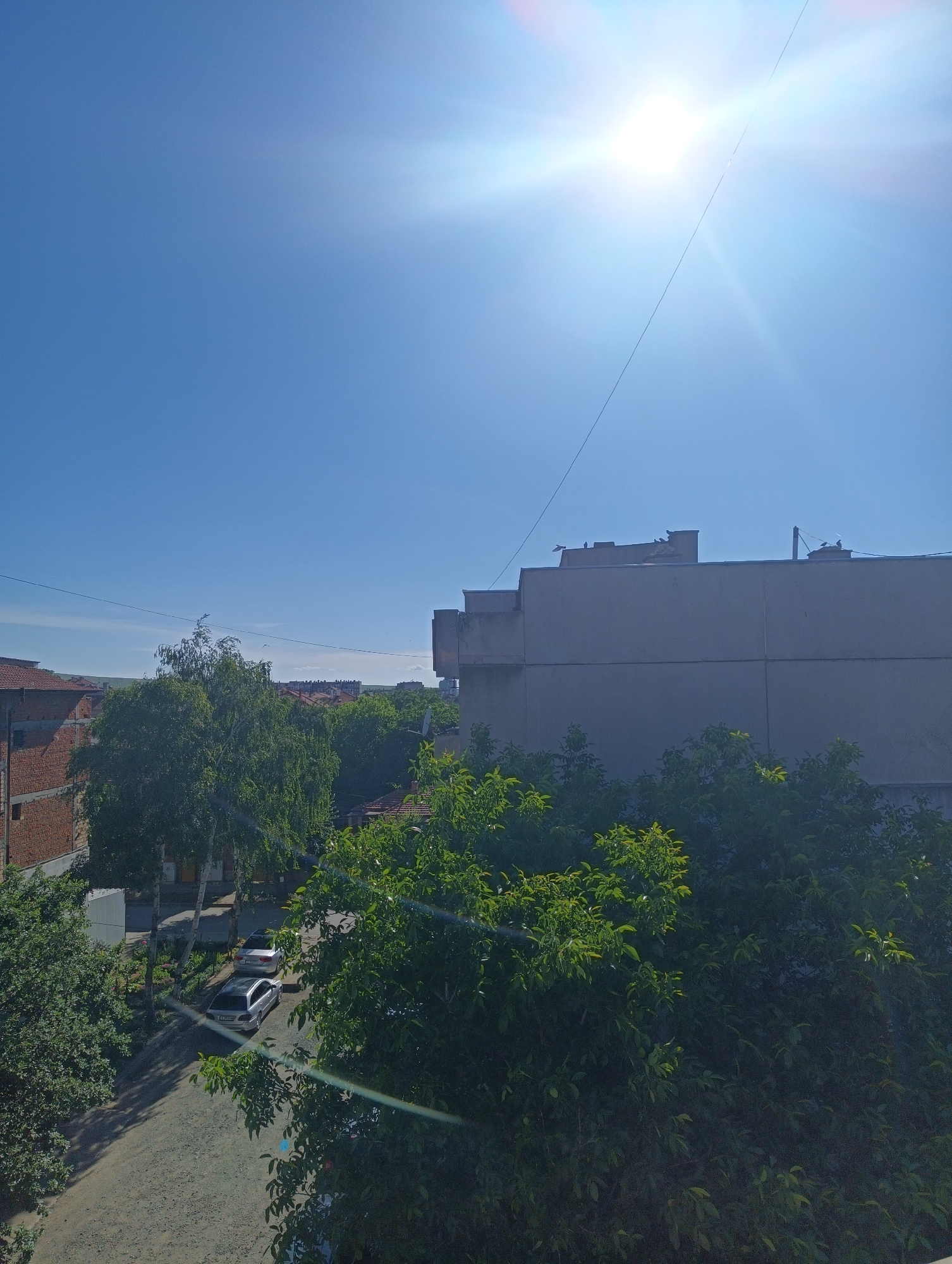 The image captures a bright, sunny day in Karnobat, Bulgaria, with the sun prominently featured in the upper right quadrant of the sky, emitting strong lens flare. The scene overlooks a residential area with a mix of buildings and lush greenery. In the foreground, dense green foliage partially obscures the view. Beyond the trees, a multi-story building with a gray facade stands out, its upper portion showing some structural elements. To the left of this building, a partially constructed brick structure is visible. Below the buildings, a gravel pathway is lined with parked cars, including a silver sedan and another lighter-colored vehicle. Several trees, including one with a birch-like trunk, line the pathway. In the distance, more buildings with terracotta roofs are visible, suggesting a typical urban landscape. The sky is a clear, vibrant blue.