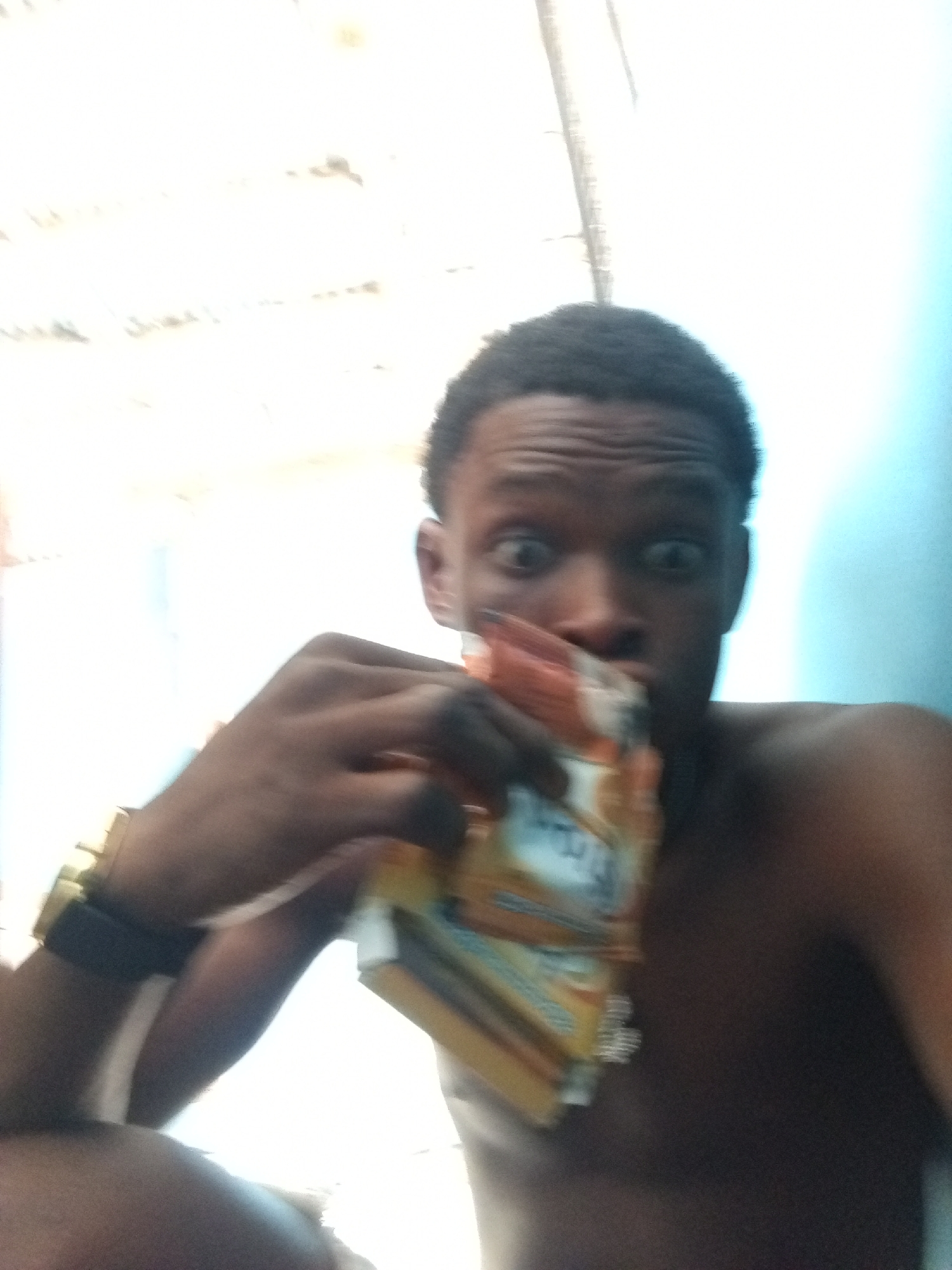 A close-up, slightly blurry, selfie-style photograph features a young Black male from the chest up. He is positioned indoors against a very bright, overexposed background, possibly a window, with a light blue wall visible on the right. The man appears shirtless, with visible shoulders and upper chest. He has short, dark hair and an expressive face, characterized by wide, open eyes and slightly furrowed eyebrows, conveying an emotion that could be surprise, playfulness, or excitement.
In his left hand, he holds a stack of small, rectangular packets, positioned in front of his mouth. These packets are primarily orange and yellow, with some white and blue elements. Although blurry, text resembling "INDOMIE" and possibly "CHICKEN FLAVOUR" can be faintly seen on the packaging, suggesting they are instant noodle packets. He wears a gold-colored watch or bracelet with a dark strap on his left wrist. The image has a high-contrast appearance due to the bright background, and the overall feel is candid and informal. The location context for this image is Ado, Nigeria.