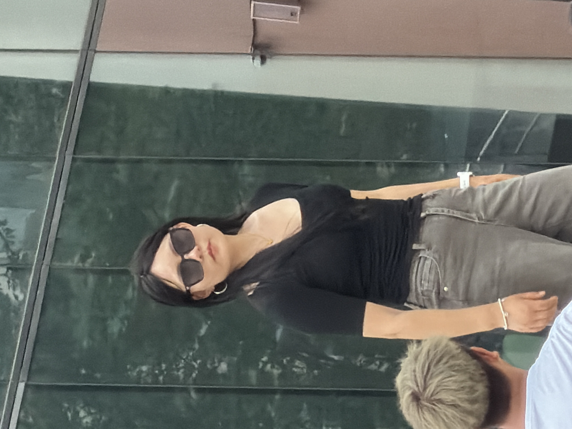 A woman with black hair and sunglasses stands in front of a glass building. She is wearing a black top and grey pants. A man with blonde hair is partially visible in the lower left corner of the image. The scene appears to be in a public urban setting in Yuzhong District, China.