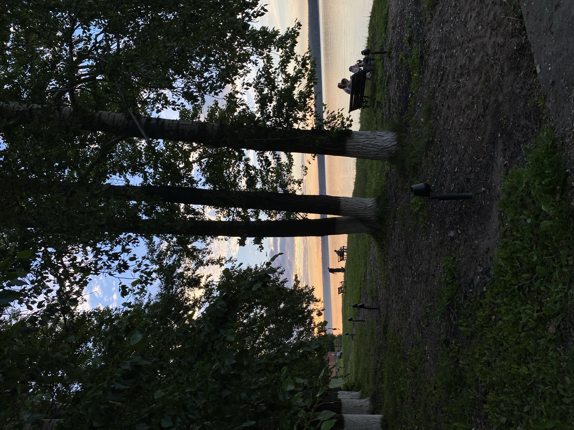 This image depicts a serene lakeside scene in Votkinsk, Russia, likely during sunset. The foreground features a grassy embankment with several trees, their trunks painted white at the base. Small, dark-colored pathway lights are intermittently placed along the edge of the grassy area.
In the middle ground, a body of water, possibly Lake Votkinsk, stretches towards the horizon, reflecting the warm hues of the setting sun. The sky above is a blend of soft blues and oranges, with scattered clouds.
Further back, along the water's edge, are several individuals. On the left, a few people are gathered near a building with a red roof. In the center, a person stands near a bench where two other figures are seated. On the right side of the image, two people are seated on a park bench facing the water.
The overall atmosphere is peaceful and tranquil, suggesting a public park or recreational area by the lake. The lighting indicates it is late afternoon or early evening.