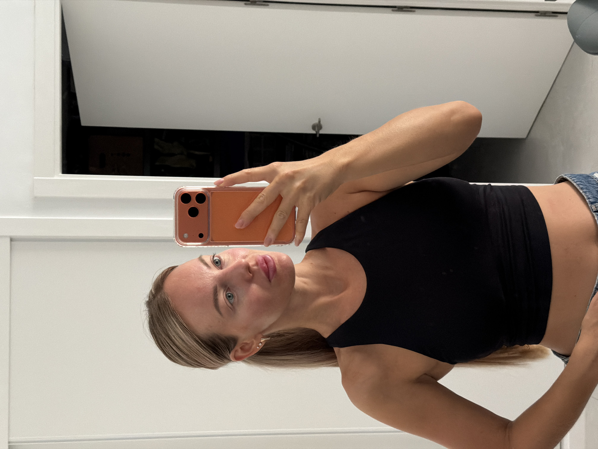 The image shows a young woman with blonde hair pulled back into a ponytail, wearing a black crop top and denim shorts. She is holding an orange-cased smartphone, taking a selfie in a mirror. The background appears to be an indoor setting with white walls and a slightly ajar door. There is no indication of the location being in Finestrat, Spain.