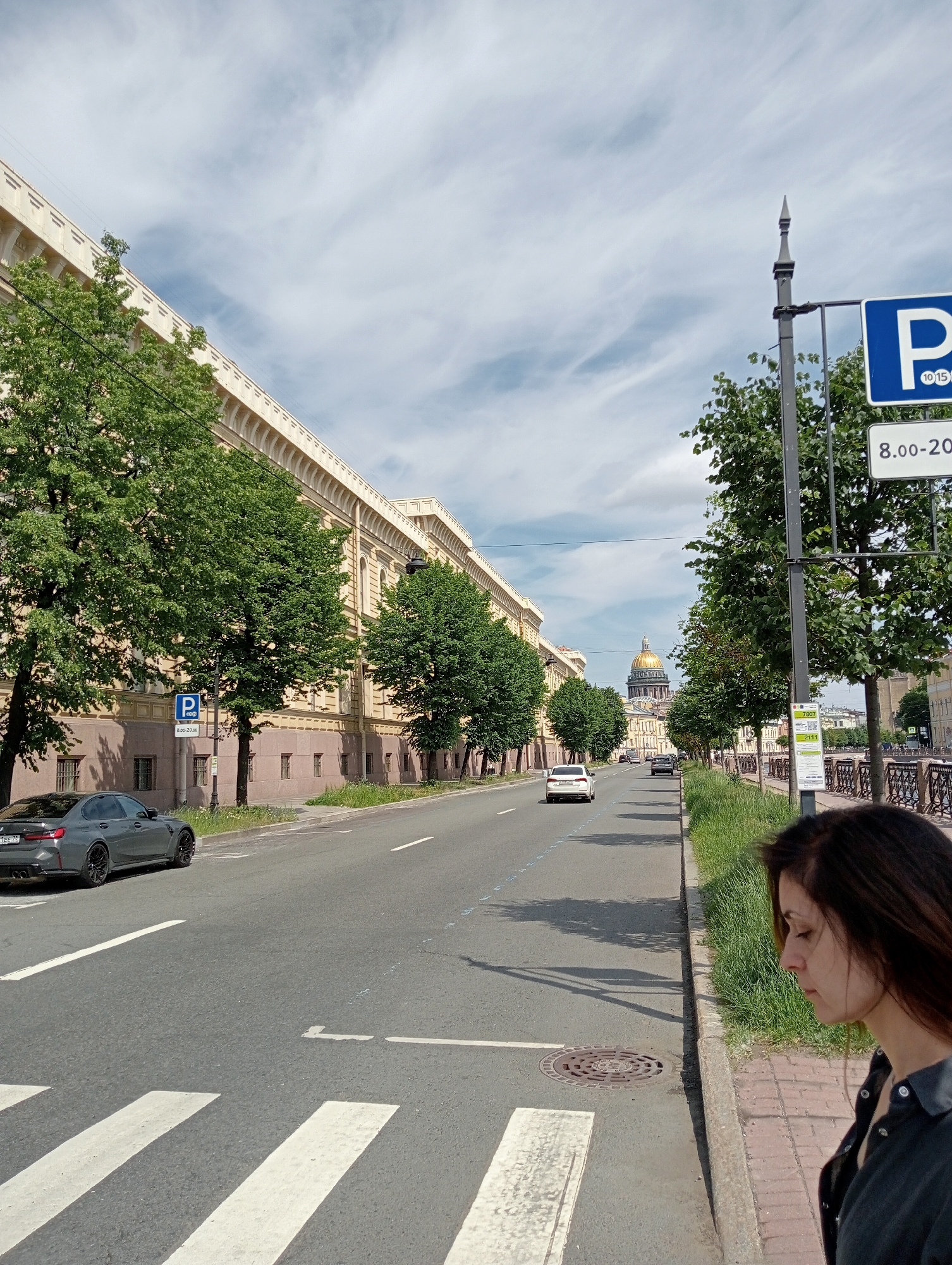 This image depicts a street scene in Saint Petersburg, Russia, likely near St. Isaac's Cathedral, which is visible in the background with its golden dome. The foreground shows a woman in profile, looking down, with her dark hair cascading over her shoulder. She is standing on a sidewalk next to a road with a pedestrian crossing. A dark grey sedan is parked on the left side of the road, and other cars are driving along the street. The architecture on the left side of the street is grand and classic, with a long, multi-story building featuring numerous windows and decorative elements. Trees line the street, casting shadows on the pavement. A parking sign is visible on the right, indicating parking restrictions from 8:00 to 20:00. The sky is partly cloudy.