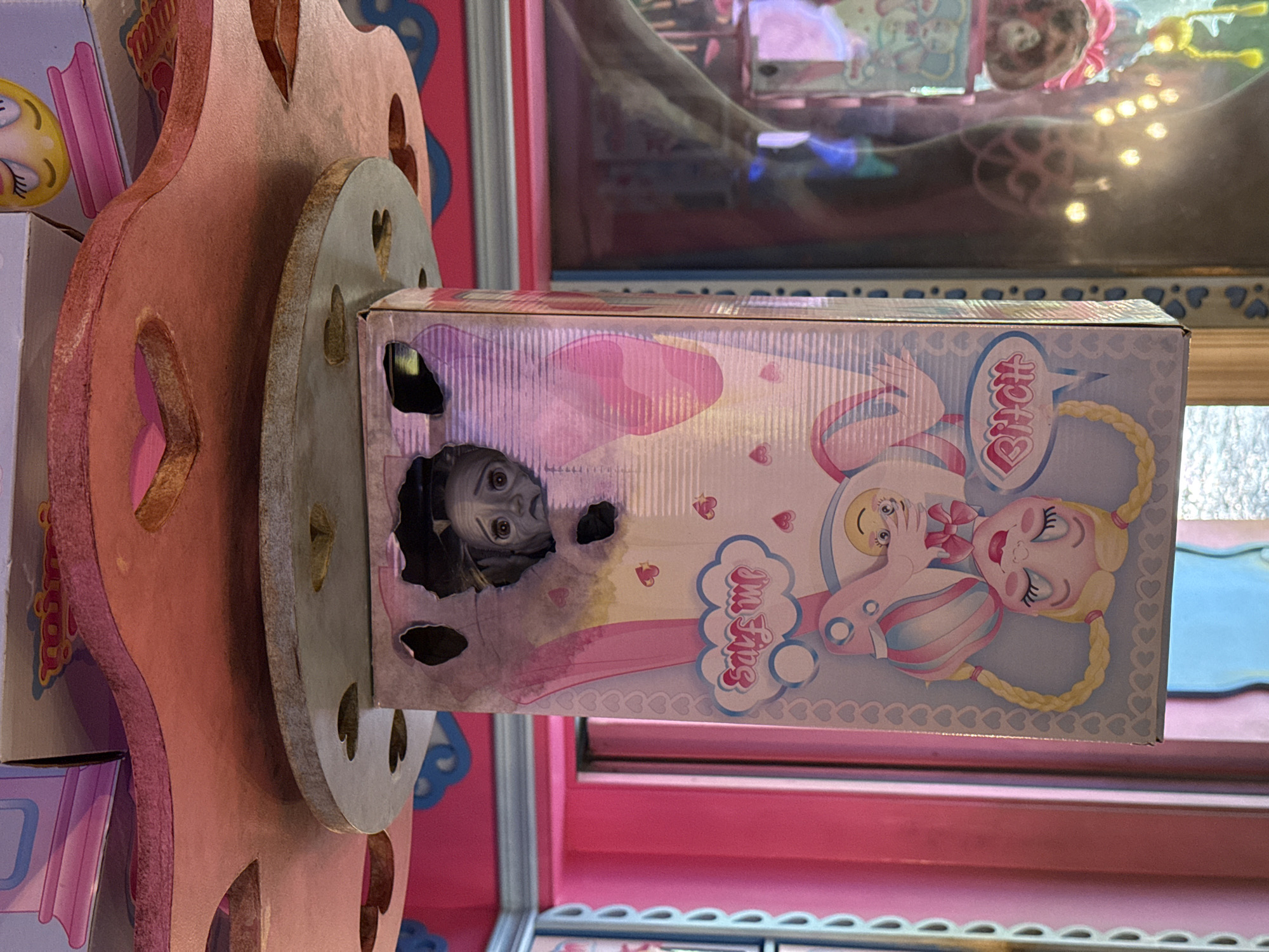 The image shows a doll box sitting on a pink and gray structure with heart-shaped cutouts. The box features an illustration of a blonde doll with braided pigtails and a baby doll. The word "Bitch" is printed in pink on the box. A doll is visible through a hole in the side of the box. The location is Unknown, Unknown.