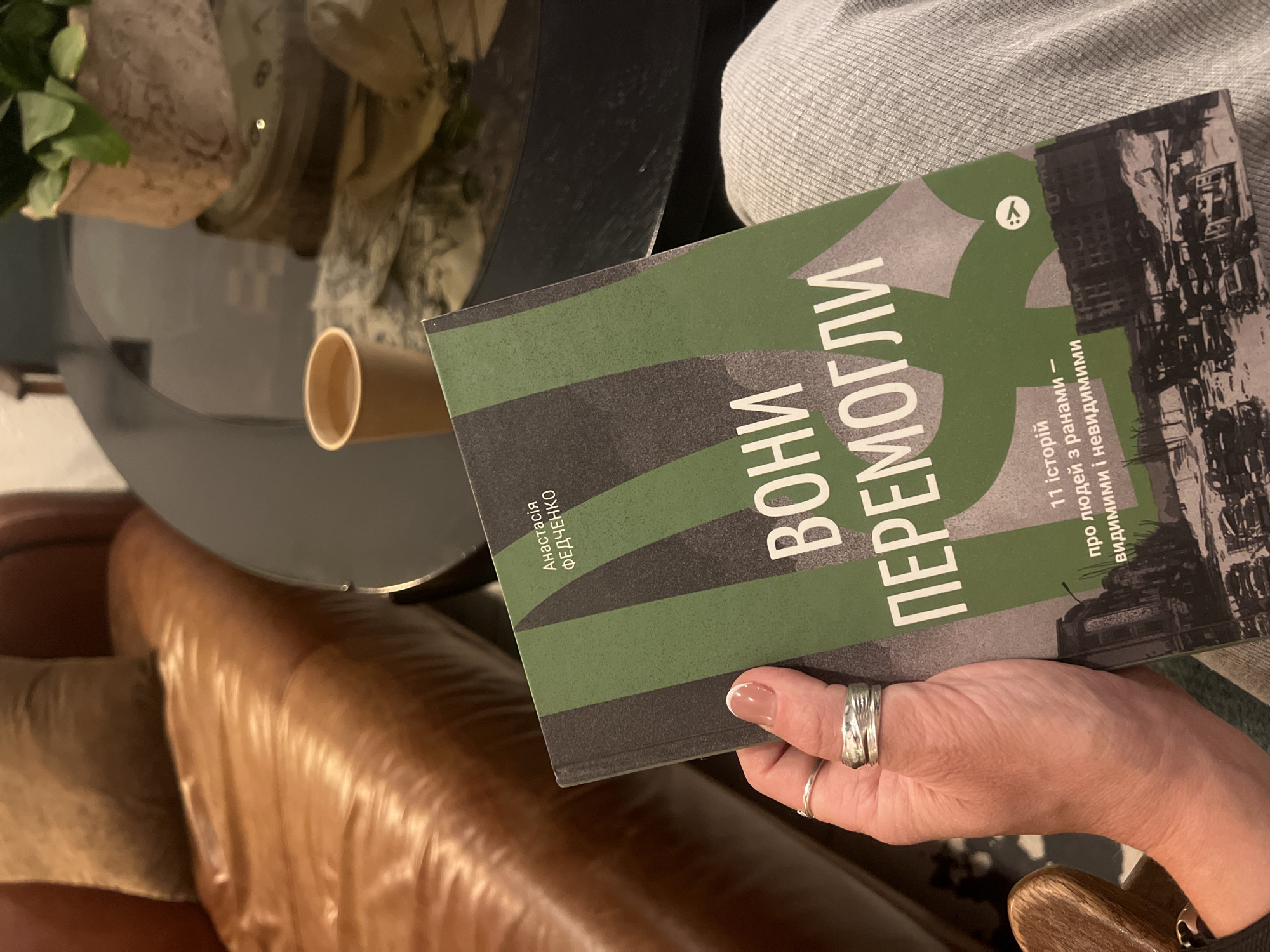 A person's hand, adorned with two silver rings, holds an open book. The book's cover features bold white text against a green and dark gray background. The text reads "ВОНИ ПЕРЕМОГЛИ" and below that, "11 історій про людей з ранами – видимими і невидимими". The background of the cover depicts a black and white image of a damaged city street with buildings and vehicles. The person is sitting on a gray, textured fabric, possibly pants or a skirt. The book is positioned over their lap. In the background, out of focus, is a glossy black round table with a paper cup of liquid and a potted plant. To the left, a portion of a brown leather sofa is visible. The setting appears to be an indoor, comfortable space. There is no explicit indication of the city of Kyiv, Ukraine, in the image itself.