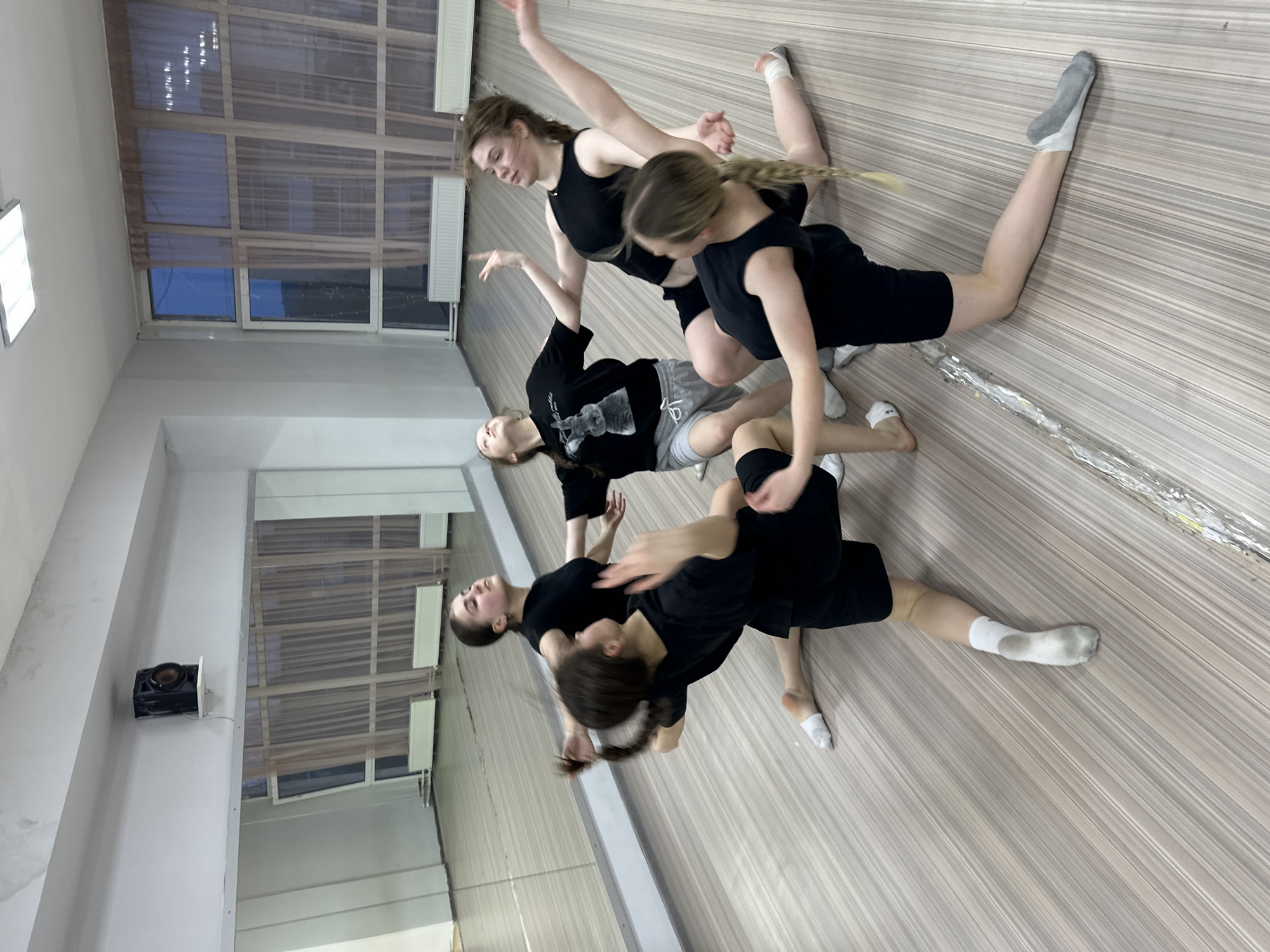 The image displays five young women engaged in a contemporary dance or movement practice within a bright studio, likely in Votkinsk, Russia. The dancers, appearing to be teenagers or young adults, are arranged in a dynamic, intertwined formation on a light-colored wooden floor. Several are lying or kneeling in fluid poses, with limbs extended and bodies angled, suggesting a moment of balance, support, and collaboration. One woman with a blonde braid kneels, reaching forward, while another in a black tank top and shorts extends her arms upwards. A dancer in a black t-shirt with an indistinct white graphic lies on her side, seemingly supported by others in the foreground who are also in various connected postures.
All participants are dressed in athletic attire, primarily black tops and shorts or light grey sweatpants. Some wear white or grey socks. Their expressions are focused and serene, reflecting concentration.
The studio features a light wooden plank floor and white walls. Large windows, partially covered by sheer, light-brown blinds, line one side of the room, providing natural light. A prominent long mirror on the opposing wall reflects parts of the room and the windows. A rectangular ceiling light fixture is visible, along with a black speaker mounted on the wall. The scene is well-lit, indicating it takes place during daytime hours. No clearly legible text is visible, although one t-shirt has a faint graphic design.