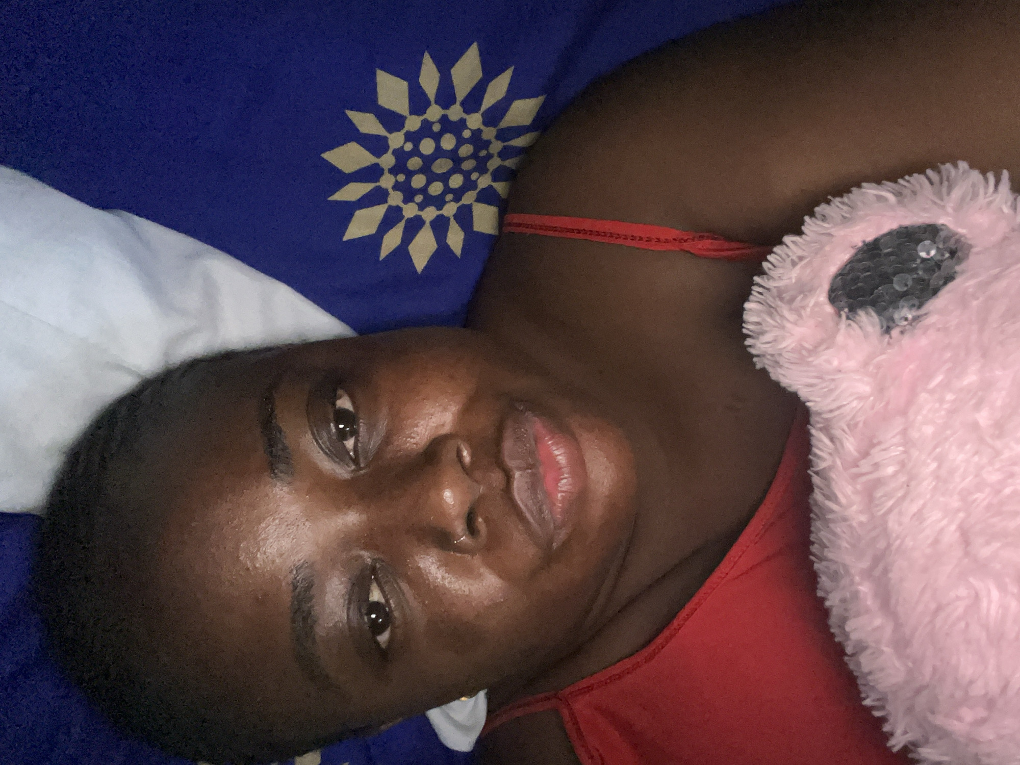 An individual with dark skin and short, dark hair is captured in a close-up, head-on shot, lying down and looking directly at the camera. The individual's face shows full lips in a slight pout and a small silver nose stud on the left nostril. The skin exhibits a natural, subtle sheen. A red top with a thin strap is visible over the right shoulder and across the chest. In the lower right of the frame, a fluffy, light pink object, possibly a stuffed animal, is held; a large, dark, circular feature resembling an eye with reflective elements is visible on it. The background consists of blue bedding adorned with a repeating light tan geometric pattern, formed by diamond shapes and central dots, and a partial white pillow or sheet in the upper left. The scene is set indoors, within a bedroom in Abuja, Nigeria.