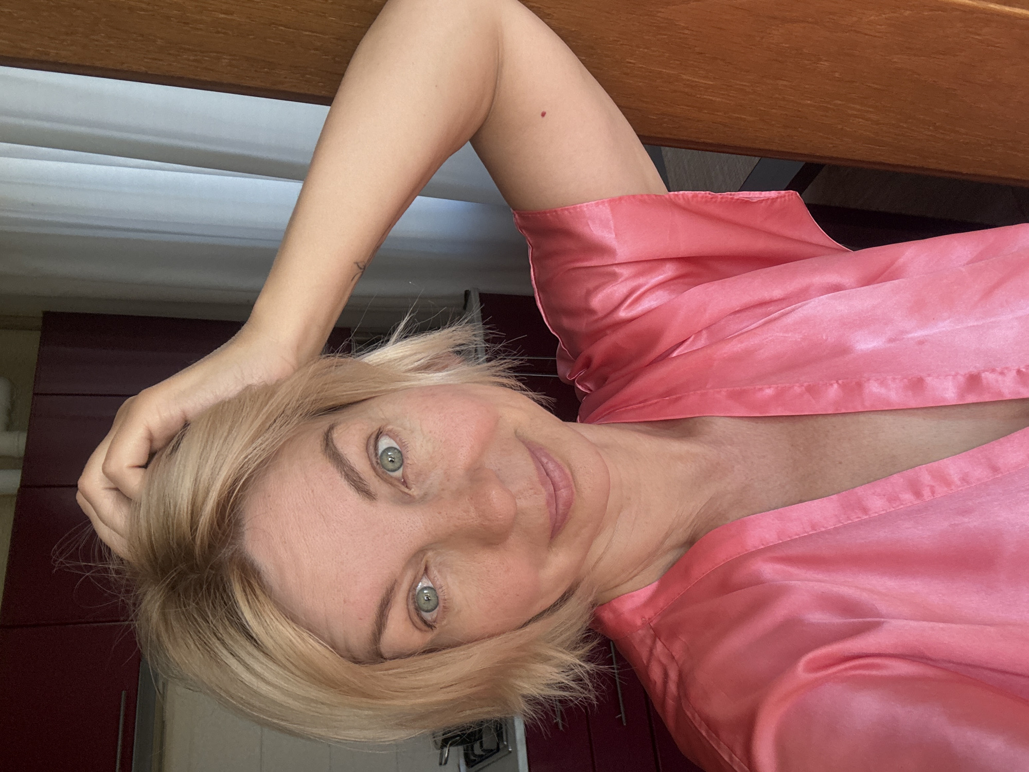 A woman with blonde, chin-length hair and green eyes is posing for a selfie indoors, wearing a pink satin robe. She is holding her right hand to her head. A faint tattoo is visible on her left upper arm. A small, dark mole is present on her right upper arm. The background includes kitchen cabinets, a curtain, and a wooden doorframe. The scene is not located in Sochi, Russia.