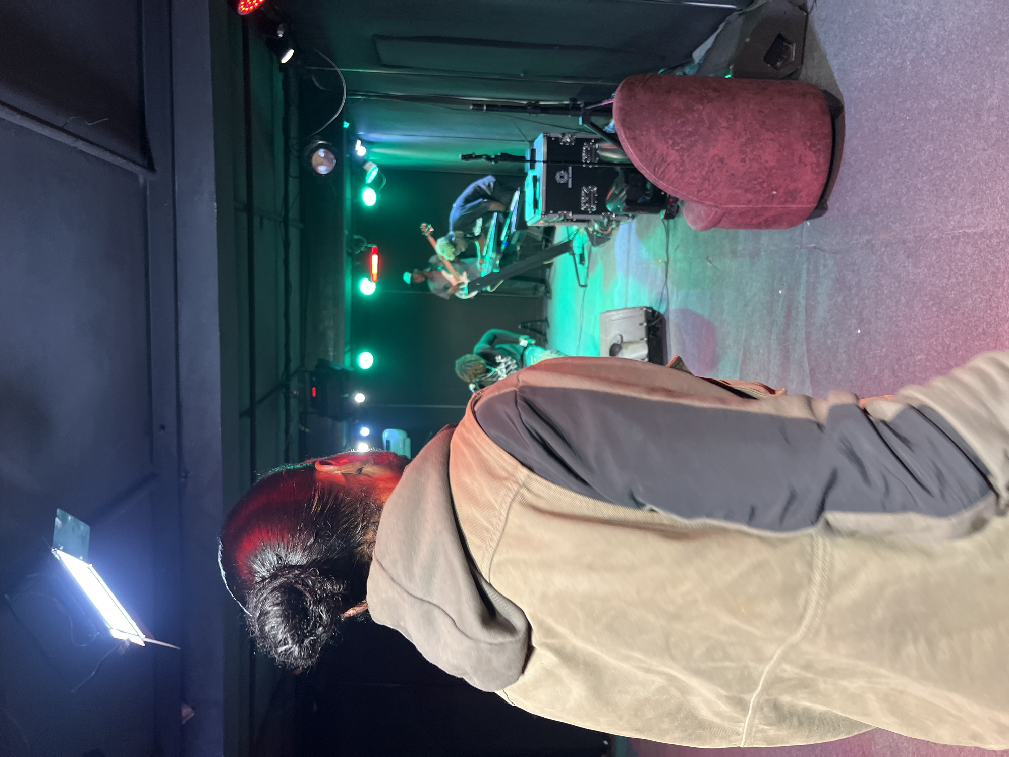 This image depicts an indoor setting, likely a performance venue or a rehearsal space. In the foreground, from a rear-facing perspective, a person with dark hair tied up in a bun is visible, wearing a brown vest over a hooded sweatshirt. The backdrop reveals a stage area with multiple individuals performing. One person is playing a bass guitar, another is seated at a keyboard, and a third is standing, possibly singing into a microphone. Stage lighting, primarily green and red, illuminates the performers and the stage floor. A red armchair is positioned to the right of the stage, and audio equipment such as speakers and a mixer are present. The overall ambiance suggests a musical performance or practice session. This scene could be located in Jos, Nigeria, where such venues host local artists and musicians.