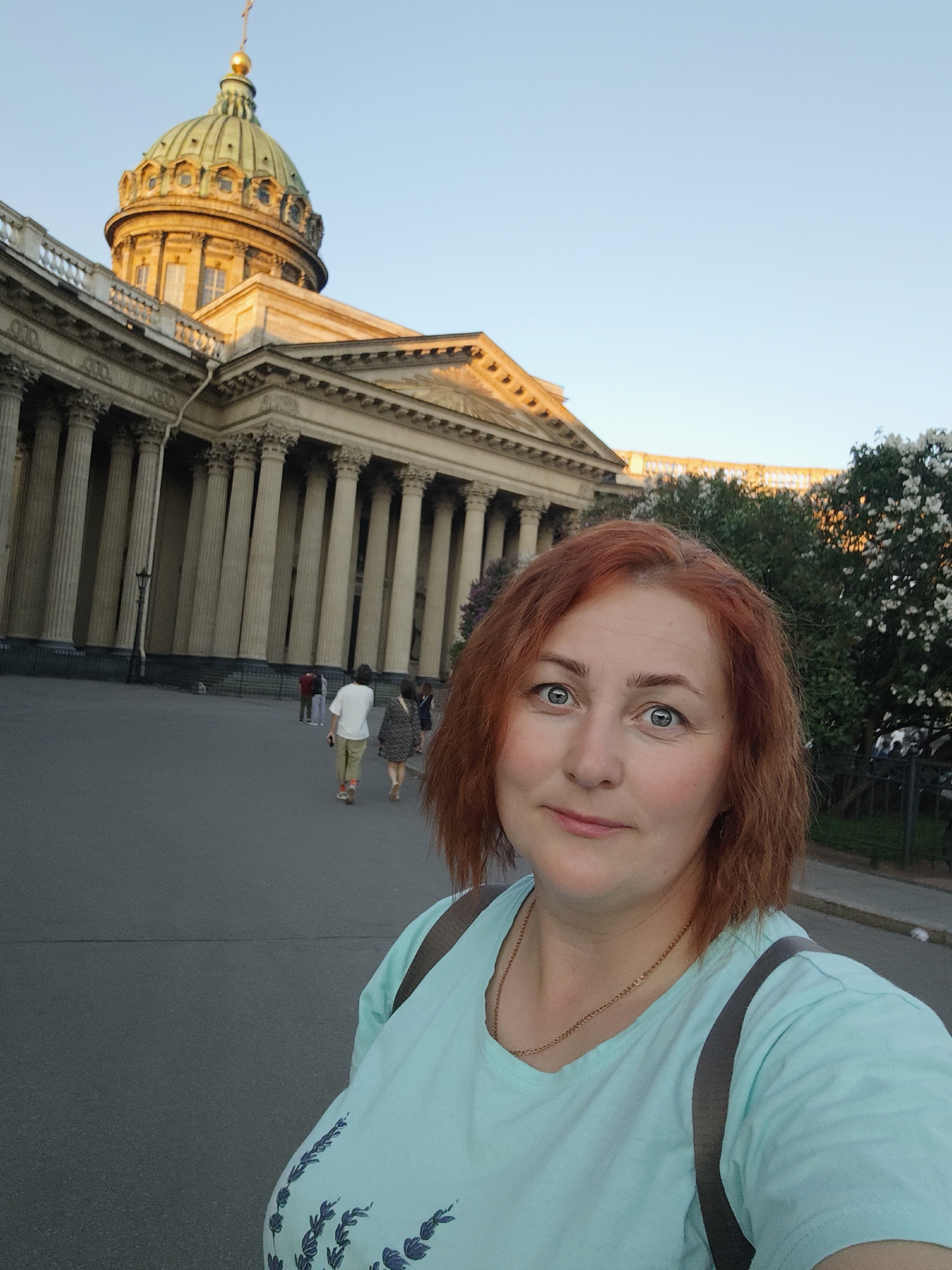 The image captures a selfie of a woman in front of the Kazan Cathedral in Saint Petersburg, Russia, during golden hour. In the foreground, a woman with reddish-brown, shoulder-length hair and blue-green eyes looks directly at the camera with a slight smile. She wears a light blue-green short-sleeved t-shirt with a darker blue leafy pattern on the lower front and a thin gold chain necklace. A dark strap from a bag or backpack is visible over her right shoulder.
Behind her, to the left, is the grand facade of the Kazan Cathedral, illuminated by golden sunlight. Its distinctive green-gold dome, topped with a golden cross, is prominent against a clear, light blue sky. The cathedral's classical colonnade with numerous tall, light-colored columns forms the backdrop.
In the mid-ground, on the paved square or street in front of the cathedral, several people are walking away from the camera. One man in a white t-shirt and light pants with orange socks walks alongside another person in a dark patterned dress. Further back, two other figures are near the cathedral entrance. To the right, a sidewalk and an area with trees bearing white blossoms and green foliage are visible. Another building with a light-colored balustrade is partially visible in the far background beyond the trees. The overall lighting suggests either sunrise or sunset.