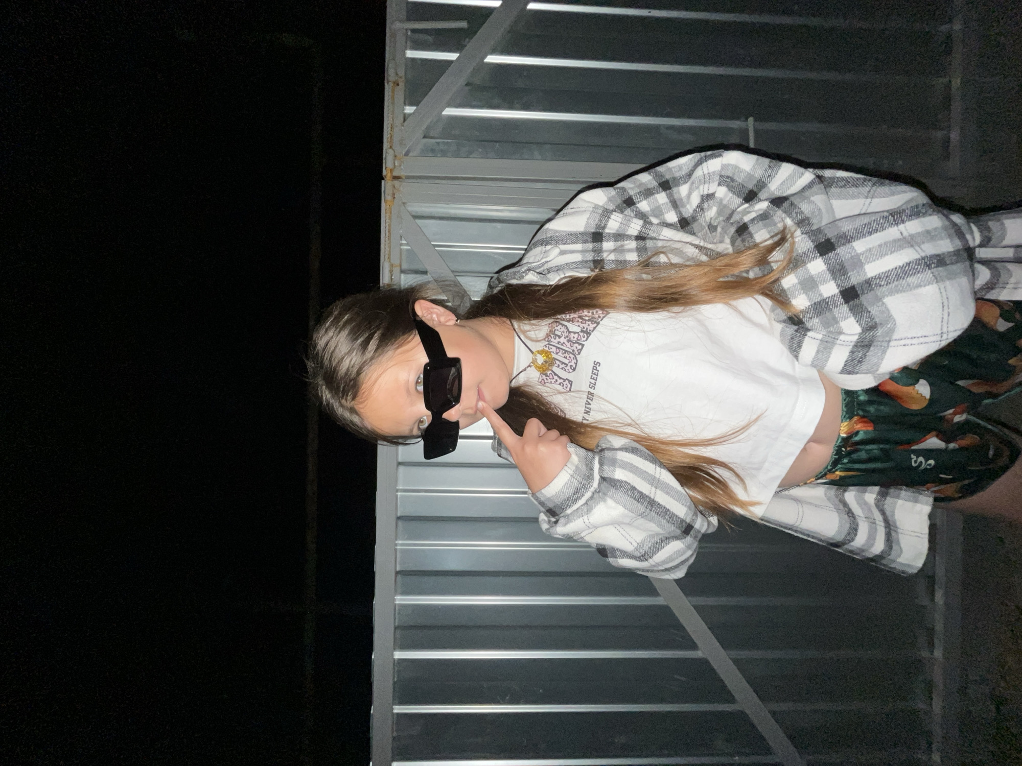 A young person stands in front of a corrugated metal wall. The person wears sunglasses, a white t-shirt, a plaid flannel shirt, and patterned pants. The location's darkness suggests a nighttime setting. A portion of the image is completely black, indicating minimal ambient light. The image's orientation is such that the subjects appear sideways. There is no indication of Novopokrovskaya, Russia.
