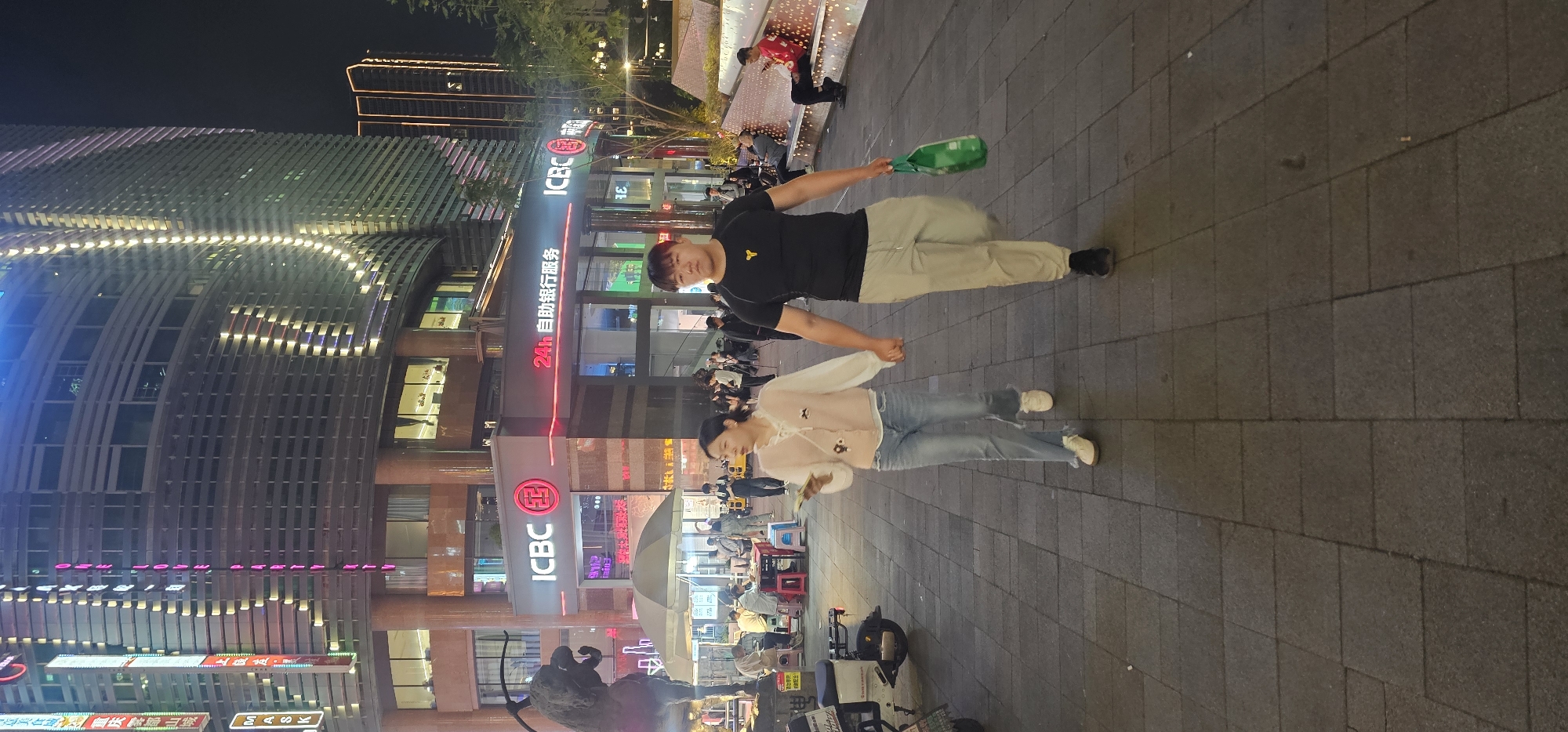 Two people walk on a stone-paved sidewalk adjacent to a busy urban street at night. The taller individual, a male, wears a black t-shirt and light-colored pants, holding a green plastic bottle in one hand. The other, presumably a female, is slightly shorter and wears a light-colored top and jeans. They appear to be walking together, possibly holding hands.
Behind them is a bustling street scene illuminated by neon lights, storefronts, and a large building identified by the ICBC logo and Chinese characters indicating a bank branch. The location is likely in Yuzhong District, China, based on the signage and architectural style. Street vendors, vehicles, and other pedestrians are visible, contributing to the vibrant urban atmosphere. A sculpture is visible nearby.