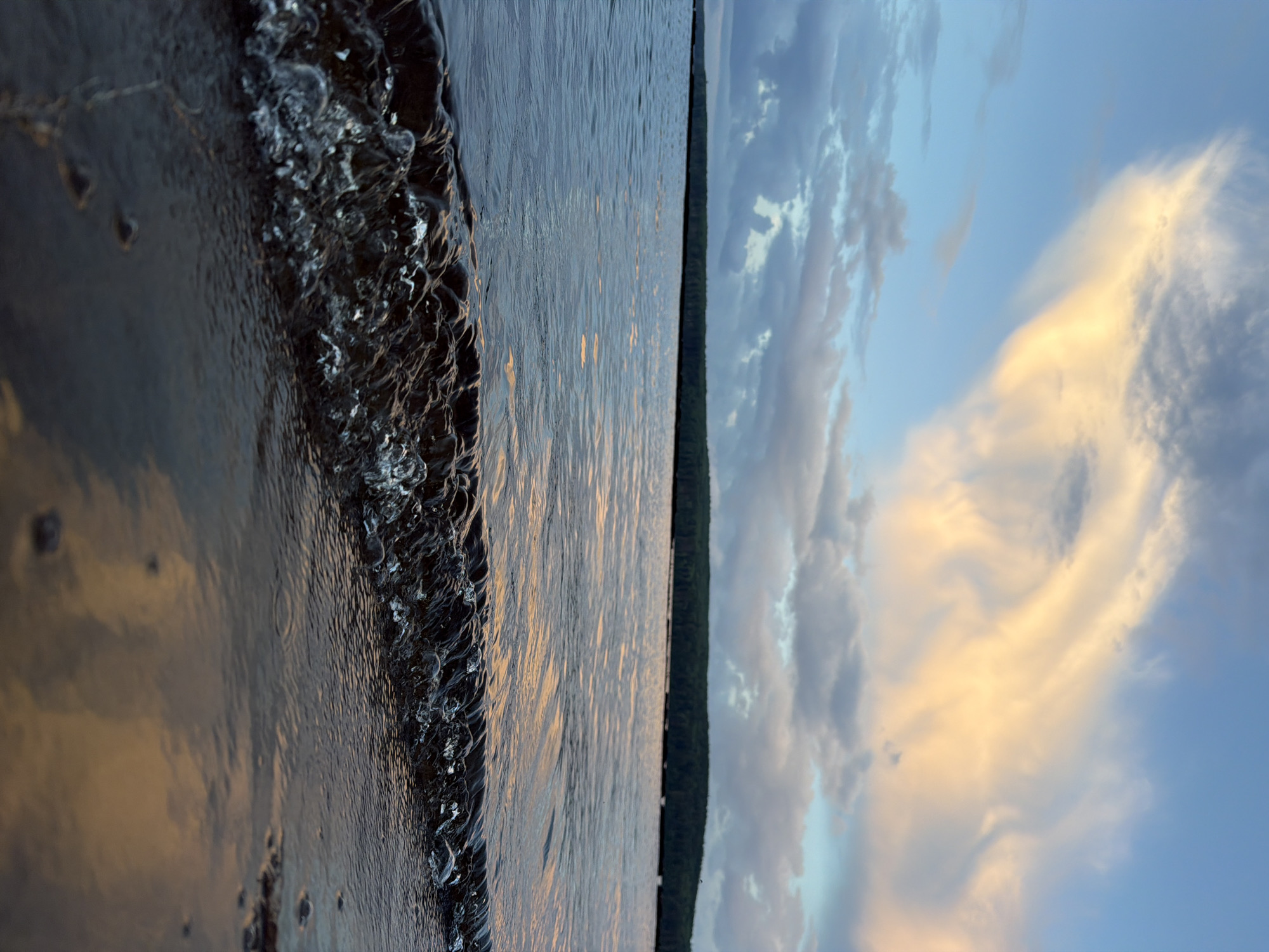 The image depicts a serene body of water, likely a lake or a wide river, with a dark, forested shoreline in the distance under a cloudy sky. The foreground features a small, translucent wave breaking onto a dark, wet shore. The sky is a blend of blue and grey clouds, with patches of soft orange and yellow light illuminating some of the clouds, suggesting it may be sunrise or sunset. The water's surface reflects the sky's colors, creating streaks of light on the rippling expanse.
While the image is aesthetically pleasing and evocative of nature, it does not contain any specific elements or context that would allow for the identification of a location within Chaikovsky, Russia. There are no recognizable landmarks, buildings, or human activity present. The scene is entirely natural, focusing on the interplay of light, water, and sky.