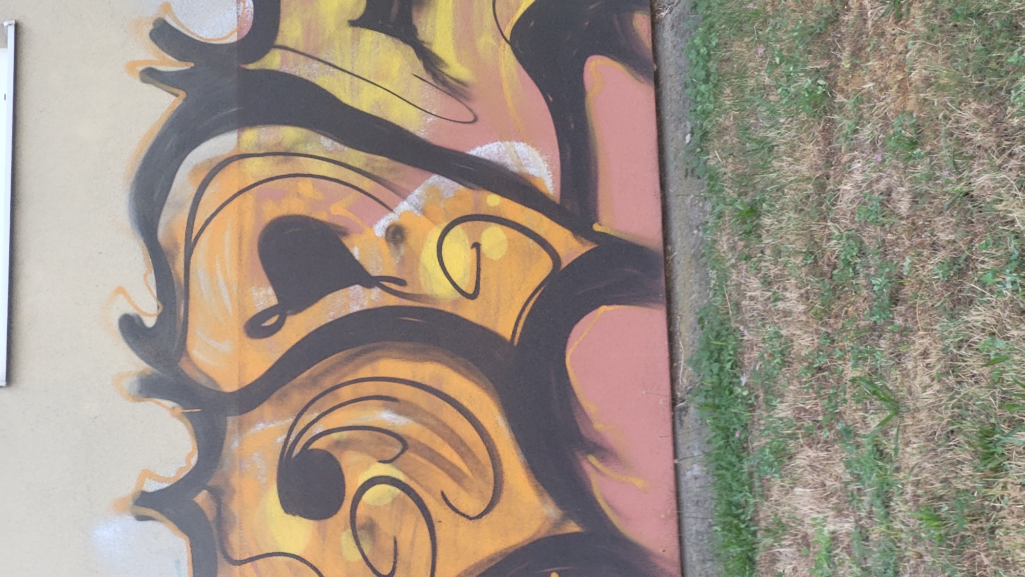 The image shows a section of graffiti on a building wall in Prešov, Slovakia. The graffiti features a stylized design with bold black outlines against a background of yellow and orange gradients. There are swirling shapes and a dark, heart-like silhouette in the center. The bottom of the wall is painted a muted pinkish-red. In front of the wall is a patch of dry, patchy grass with some green weeds growing through it. The overall scene suggests an outdoor urban environment.