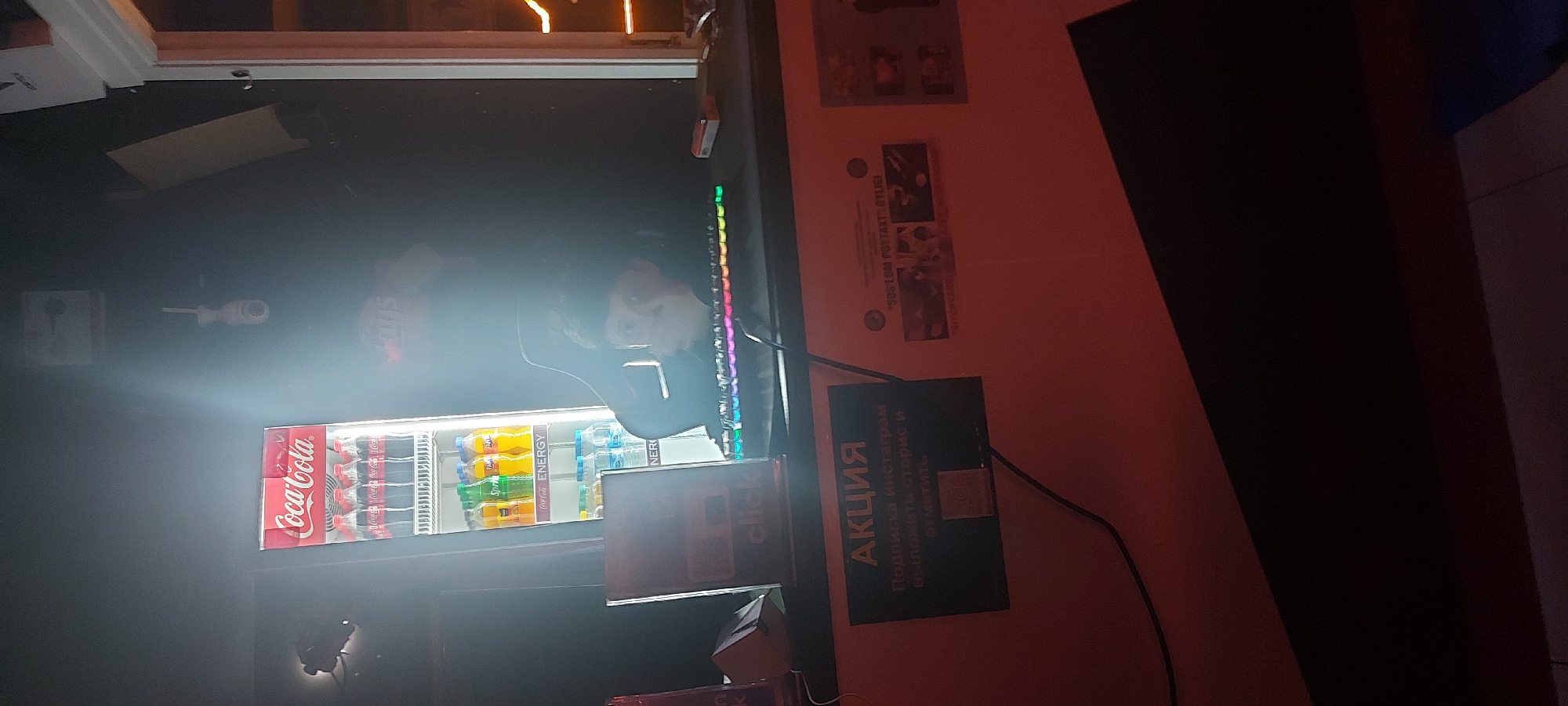 The image shows a man's reflection in a brightly lit Coca-Cola vending machine. The machine contains rows of Coca-Cola, Fanta, Sprite, and energy drinks. Next to the machine is a black sign with white text in Cyrillic reading "АКЦИЯ Подписка инстаграм Выложить сторис и отметить", advertising a promotion to subscribe to Instagram and post a story. To the right of the sign is another sign with the Cyrillic text "SOS LOM POYTAXT OYL" and a photo, and above that are some small posters. The vending machine and signage suggest a commercial environment, possibly a shop or entertainment venue. The presence of Cyrillic text indicates a location in a country where Russian is spoken, which could be the city of Tashkent, Uzbekistan, as specified in the prompt. There is a keyboard and mouse sitting on top of the vending machine. A black cabinet or wall is to the right of the wall with the posters.