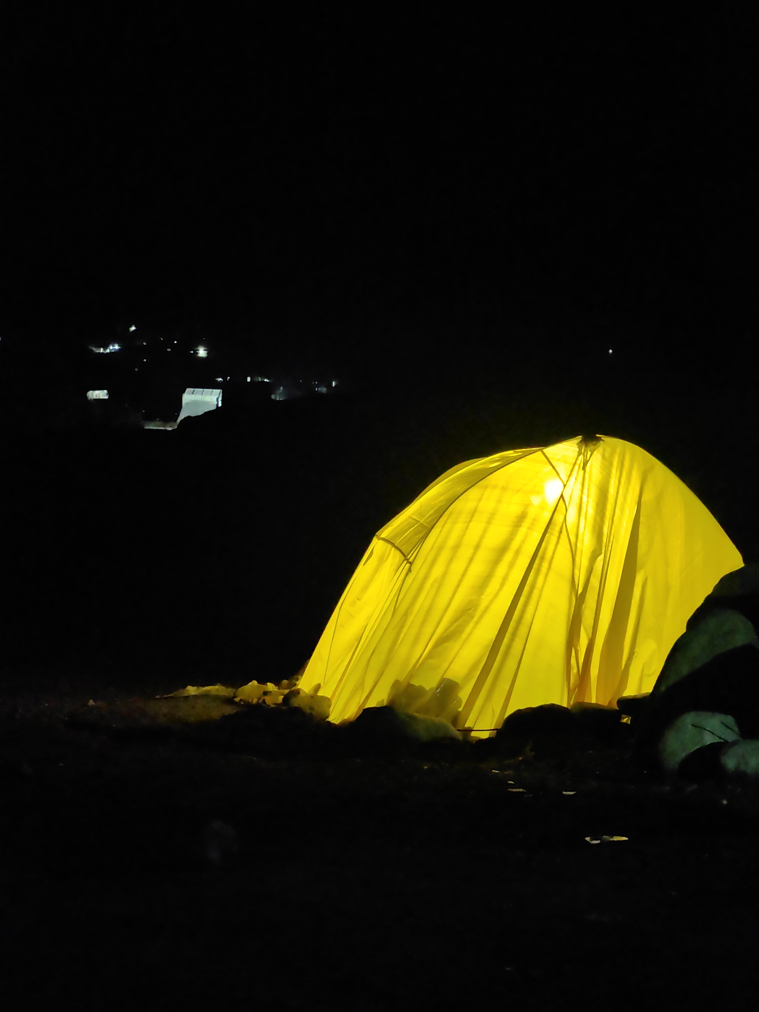 A brightly lit yellow tent is set up at night in the foreground, casting a warm glow. The tent appears to be pitched on uneven, dark terrain, possibly rocky or sandy. Behind the tent, in the distance, faint lights suggest a settlement or town, consistent with the location of Palchan, India. The sky is dark and starless. The overall scene evokes a sense of camping or an outdoor overnight stay in a remote area.