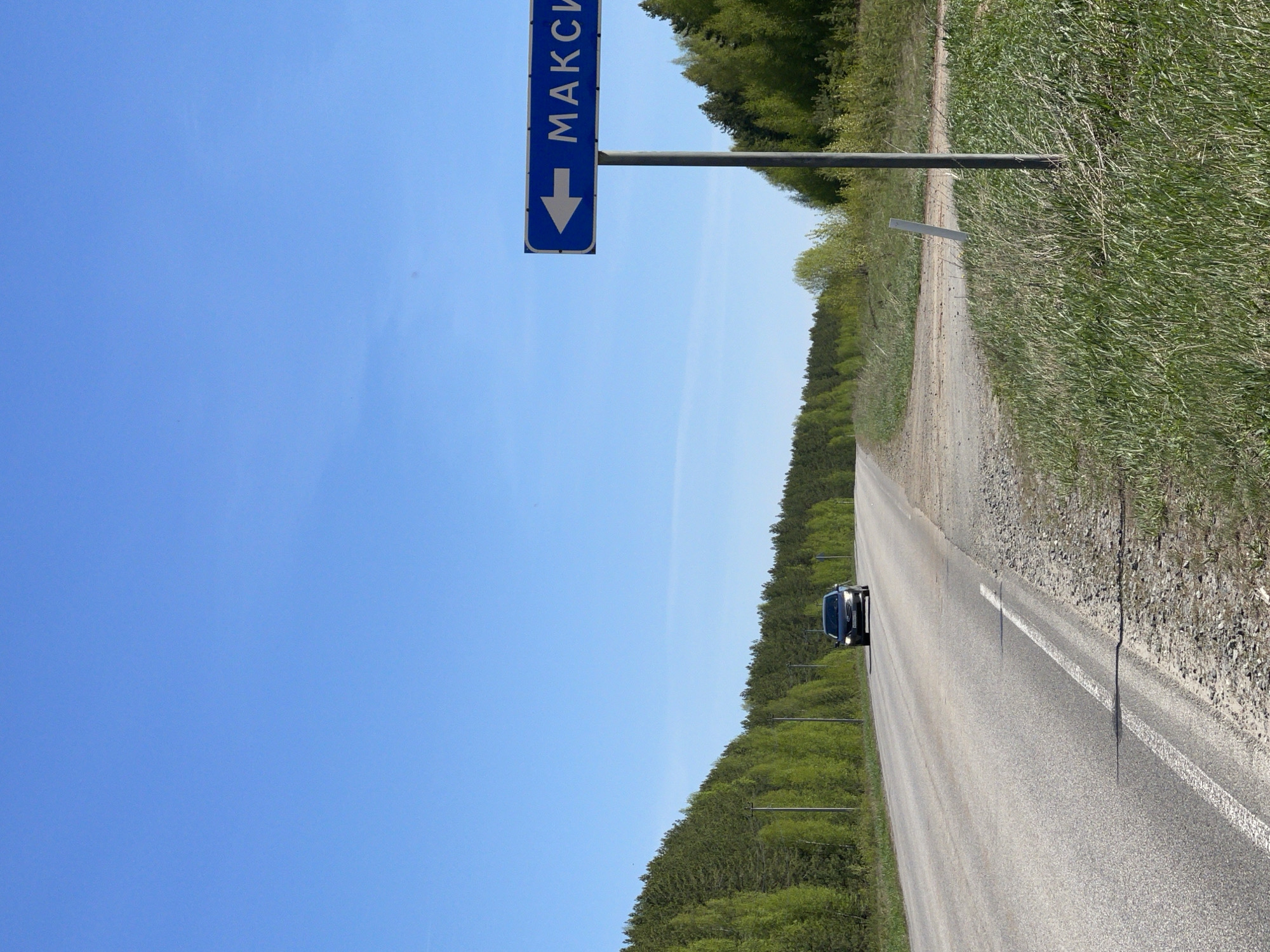 The image shows a straight asphalt road in Перевозное, Russia under a clear blue sky. A dark-colored car is traveling on the road, which has a white center line. A blue sign with a downward-pointing arrow and the Cyrillic word "МАКСИ" (MAKSI) is visible on the left side of the road. Both sides of the road are lined with green vegetation, including trees and tall grass. The ground on the side of the road is covered in gravel and debris.