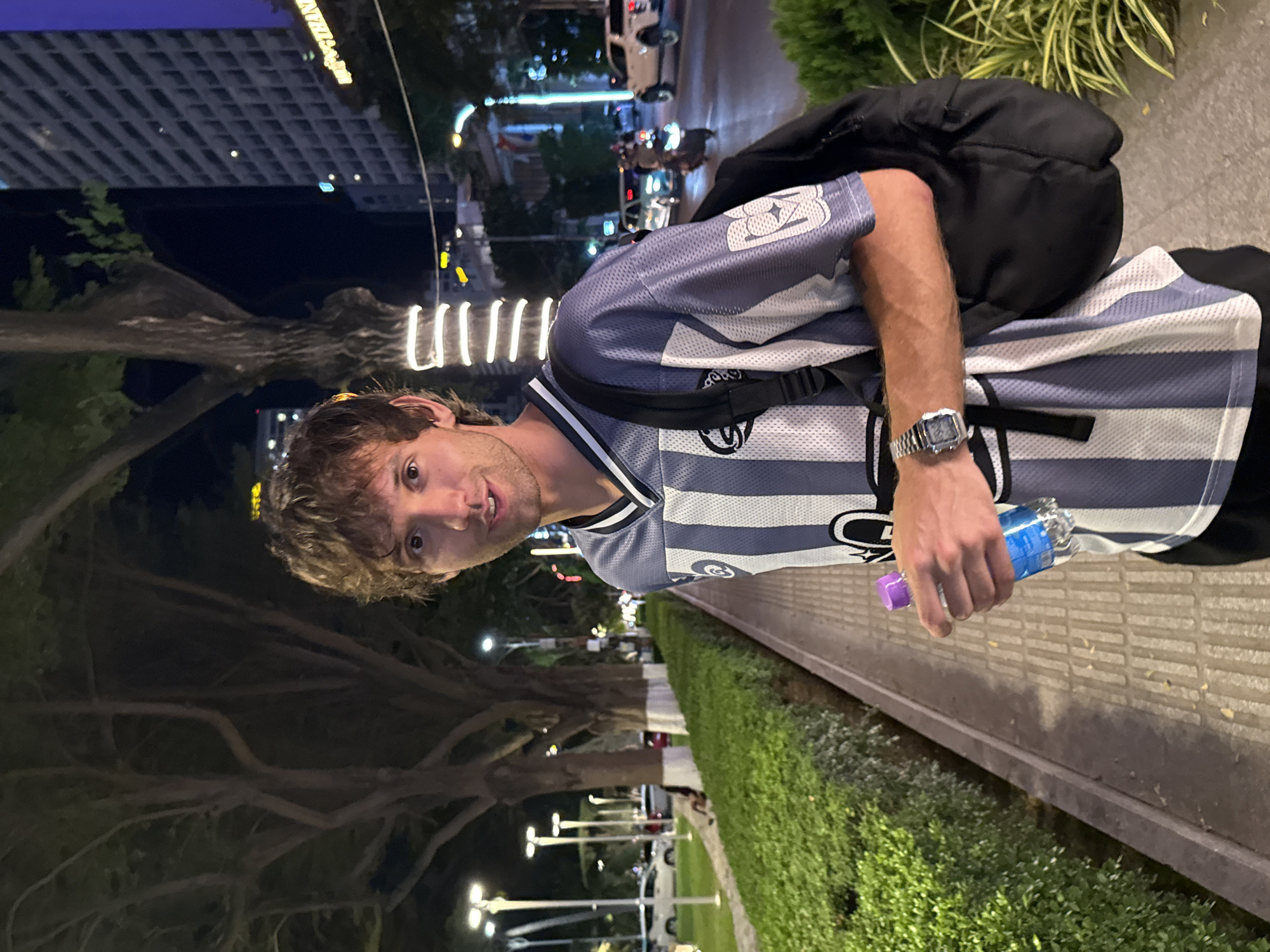A man stands on a paved walkway at night in Nha Trang Ward, Vietnam. He is wearing a striped blue and white jersey with the number 23 on the sleeve, a black backpack, and a silver wristwatch. He holds a plastic water bottle in his right hand. Behind him is a row of manicured green bushes. In the background, city lights, buildings, and traffic are visible. A large tree with winding branches is to his left.
