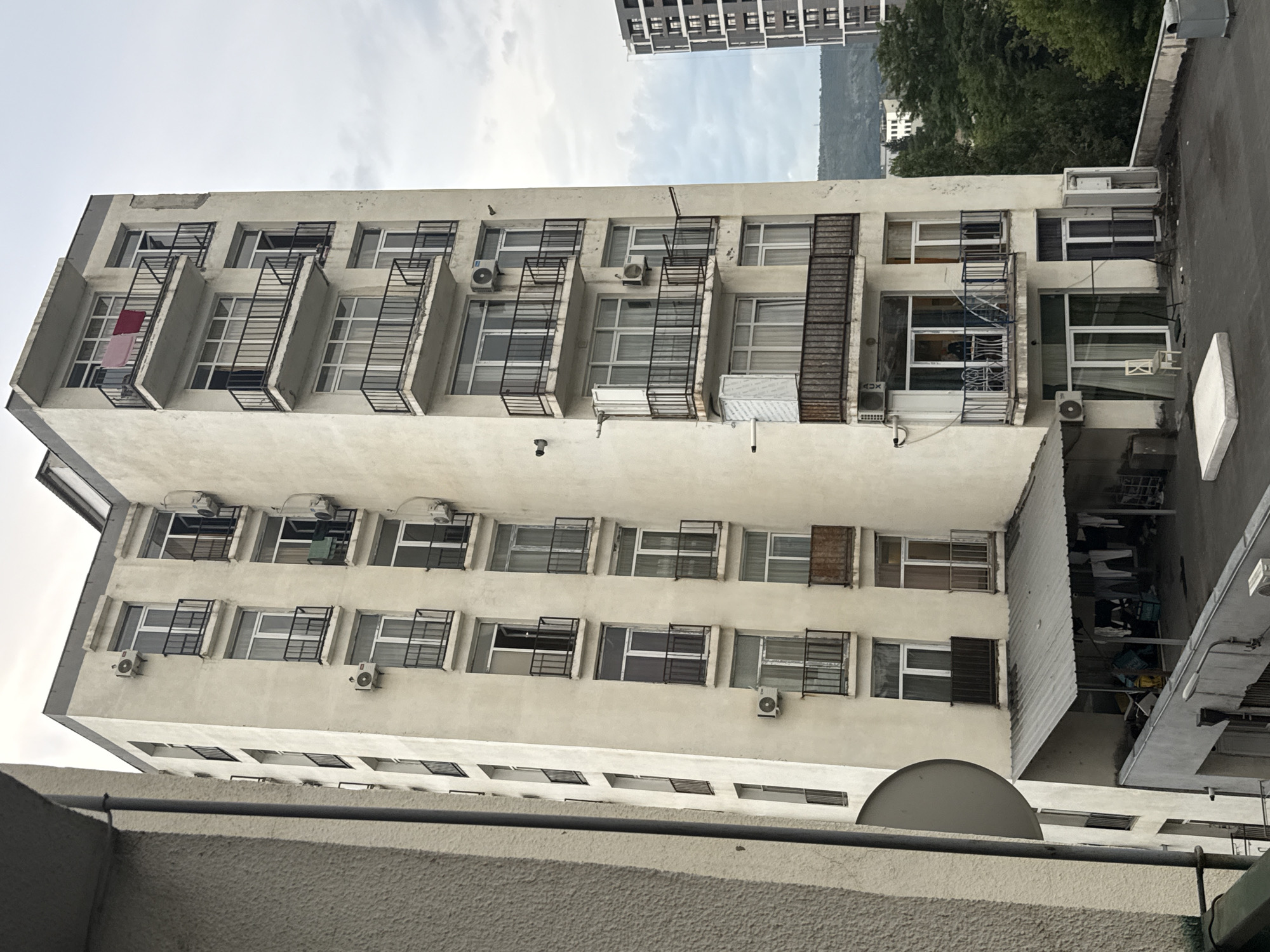 The image captures an exterior view of a tall, multi-story apartment building with a pale yellow facade, likely situated in Tbilisi, Georgia. The building features numerous balconies with black metal railings, and many windows and air conditioning units are visible. Some balconies have clothes drying, and one has two chairs and a table. At the base of the building, there's a covered area with laundry hanging and various items stored. A mattress rests on the pavement in the foreground. To the right, a more modern, dark-colored building is partially visible, and beyond the buildings, a lush green hillside is seen under an overcast sky.