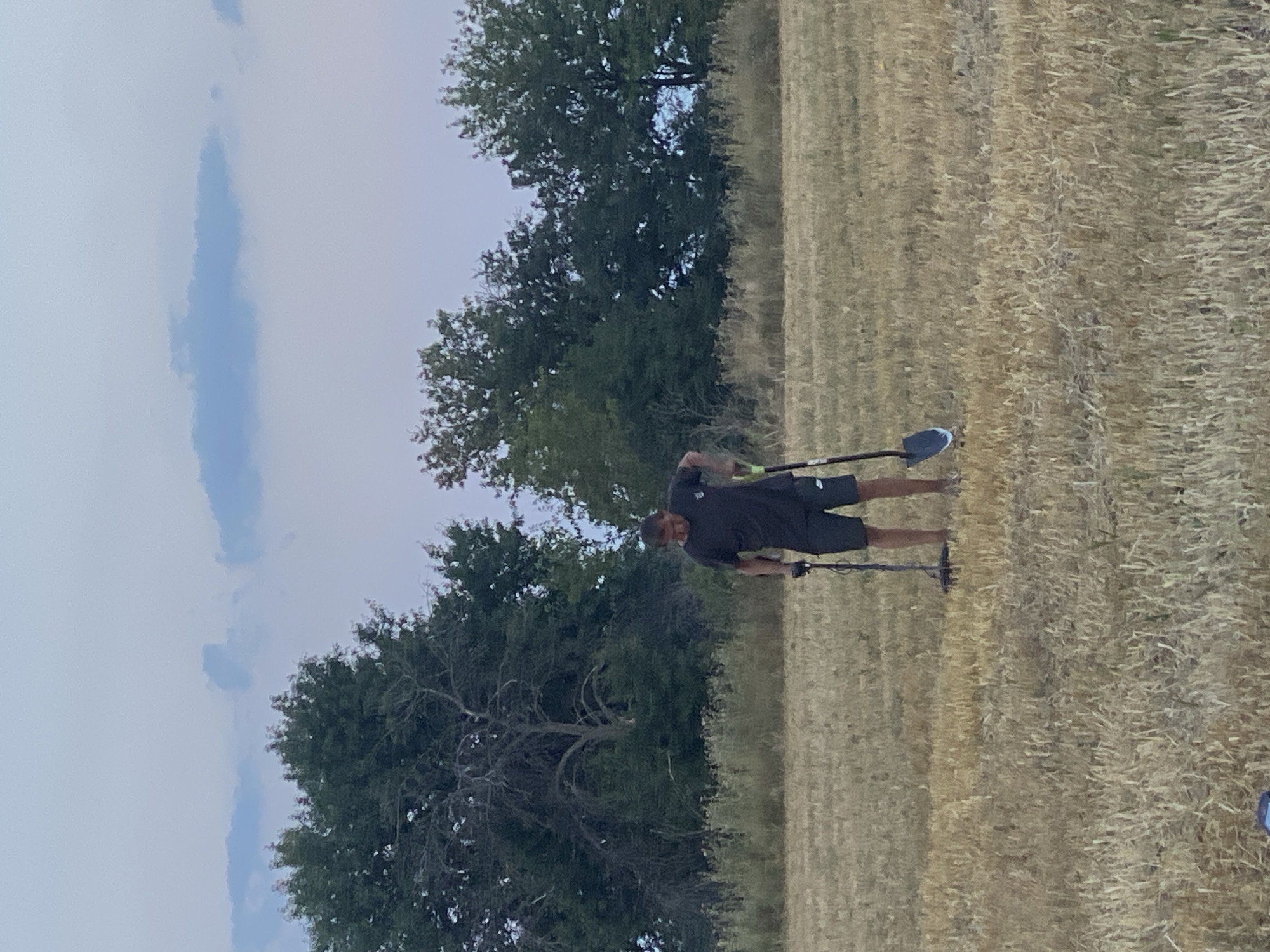 A man is metal detecting in a field of dry grass. He is wearing a black t-shirt and shorts. He is holding a metal detector in his left hand and a shovel in his right hand. The field is surrounded by trees. The sky is overcast. The scene appears to be in the city of Hoholeve, Ukraine.