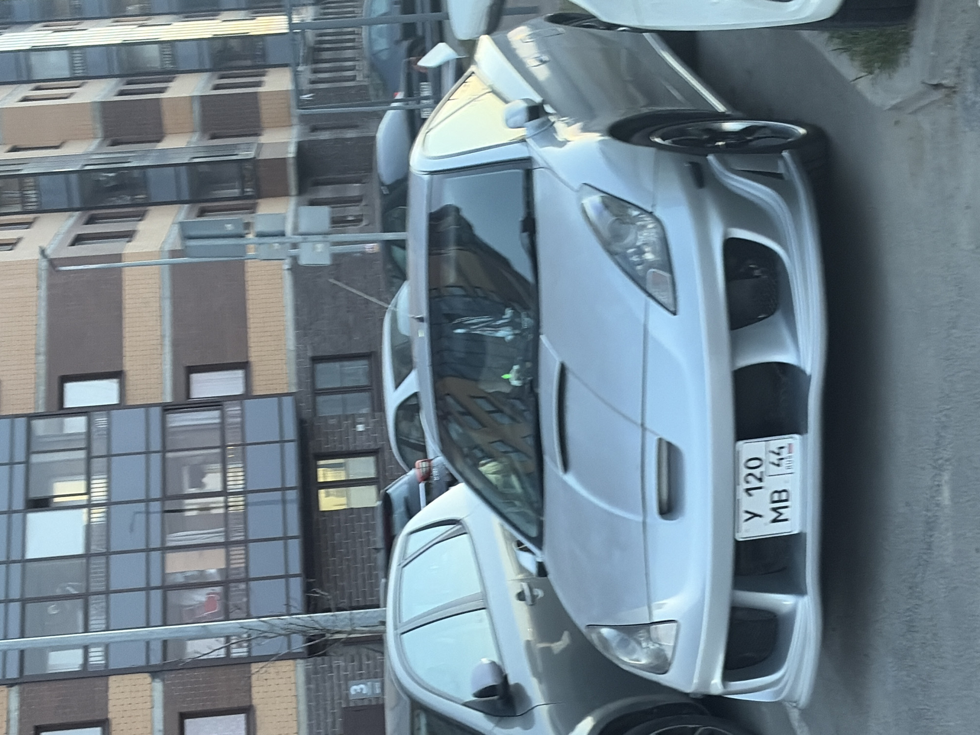 The image shows a silver Toyota Celica parked on a street in Saint Petersburg, Russia. The car has a modified front bumper and a license plate that reads "Y 120 MB 44". A multi-story residential building with brown and gray facades is in the background. There is a traffic light visible on the street. The environment appears urban. Other cars are parked nearby.