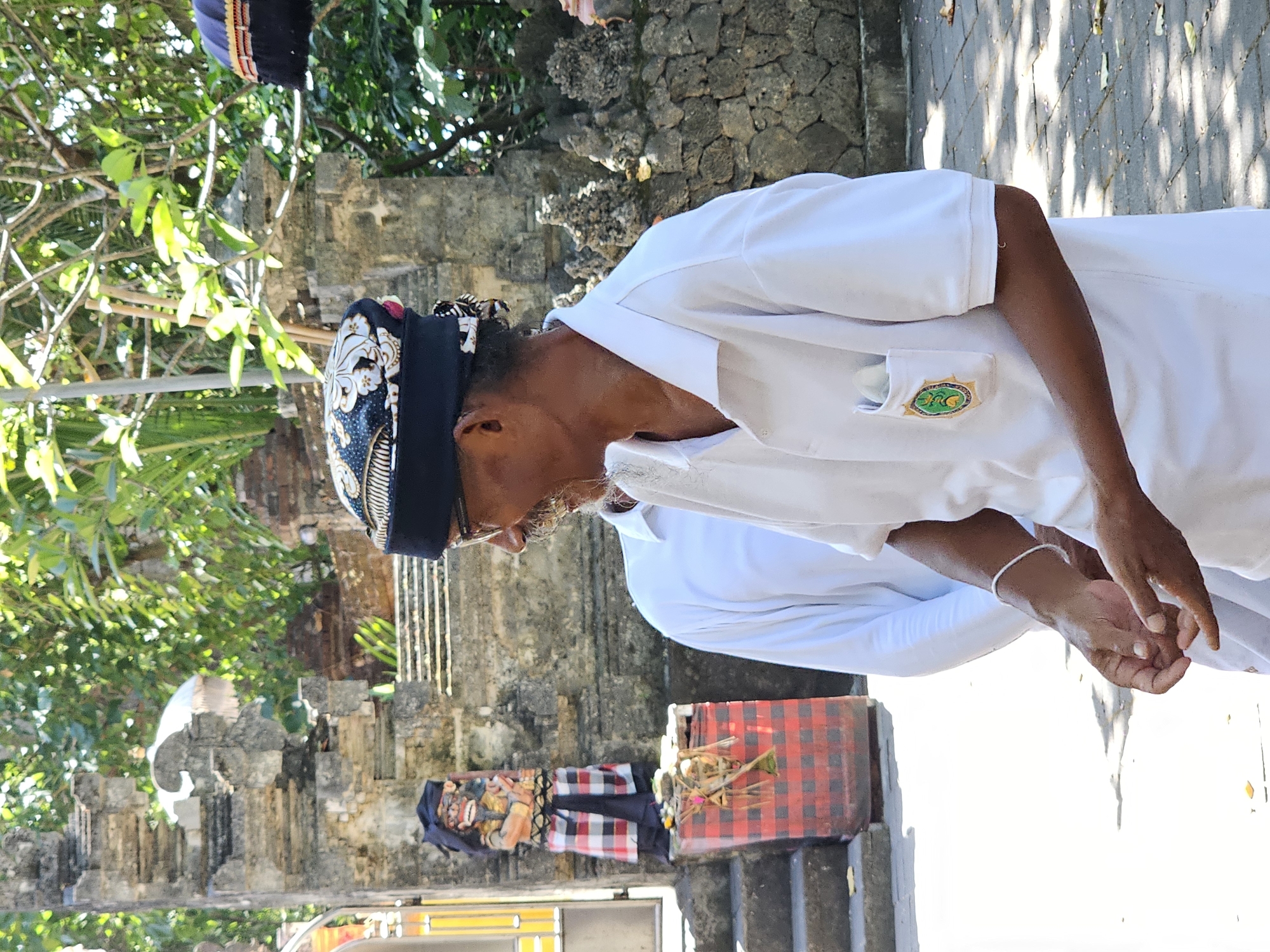 The image captures a scene in Denpasar, Indonesia, likely at a traditional Balinese temple or ceremonial site. In the foreground, an adult male with a goatee and glasses, wearing a white polo shirt and a black and gold patterned head covering, is looking to his right. His hands are clasped in front of him, and he wears a white bracelet on his left wrist. A small, circular emblem with green and gold accents is visible on the left breast pocket of his shirt. To his left, another person in a white long-sleeved shirt is partially visible. In the background, a stone structure resembling a temple gate with intricate carvings stands. To the left of the gate, a wooden effigy dressed in a red and black checkered sarong and a dark headdress is positioned on a red and black checkered base. The environment is outdoors, with sunlight casting shadows on a paved ground, and lush green foliage is visible in the upper portion of the frame.