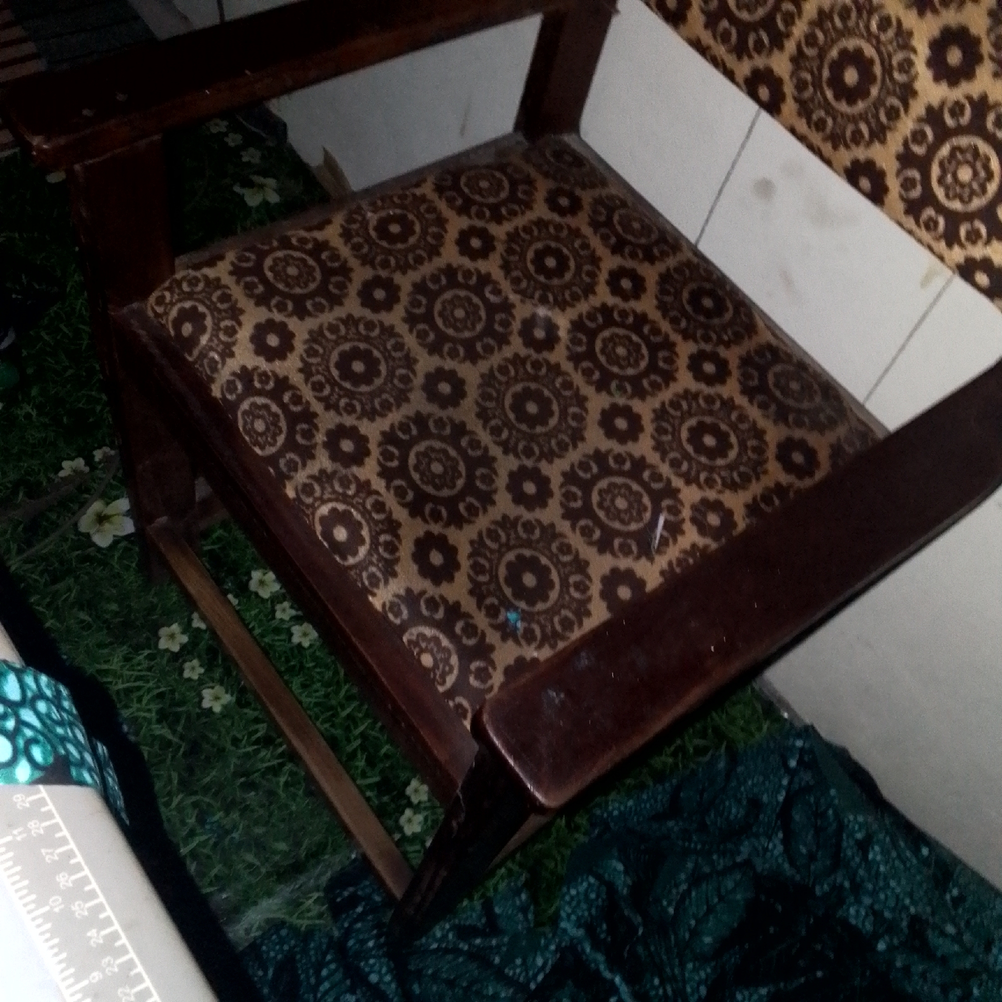 An overhead shot shows a dark brown wooden armchair with a patterned seat and back. The seat is upholstered in fabric with a repeating pattern of dark brown floral motifs on a tan background. The armchair has visible wear and tear, with scuff marks and scratches on the wooden arms and legs. The chair is placed on a dark green fabric with a subtle floral pattern. A white object with black markings and numbers, resembling a ruler or measuring tape, is visible to the left of the armchair, partially covering the patterned seat fabric. The background appears to be a wall and a wall-mounted object with a similar floral pattern as the seat. A green, patterned fabric is draped over the floor to the right of the chair. The scene appears to be indoors.