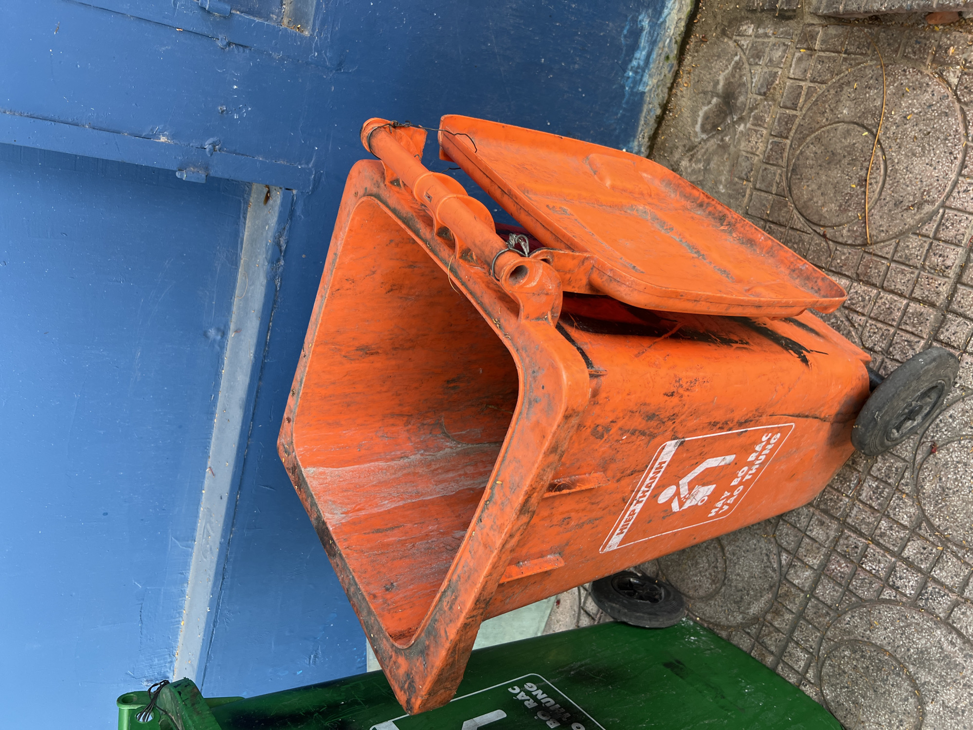 An orange, wheeled trash bin with a partially open lid sits on a concrete sidewalk in front of a blue wall. A green trash bin is partially visible to the left of the orange bin. The orange bin has a white sticker with a graphic of a person disposing of waste and text in Vietnamese. The scene appears to be outdoors in Nha Trang, Vietnam, based on the text on the bin. The ground is paved with square concrete tiles.