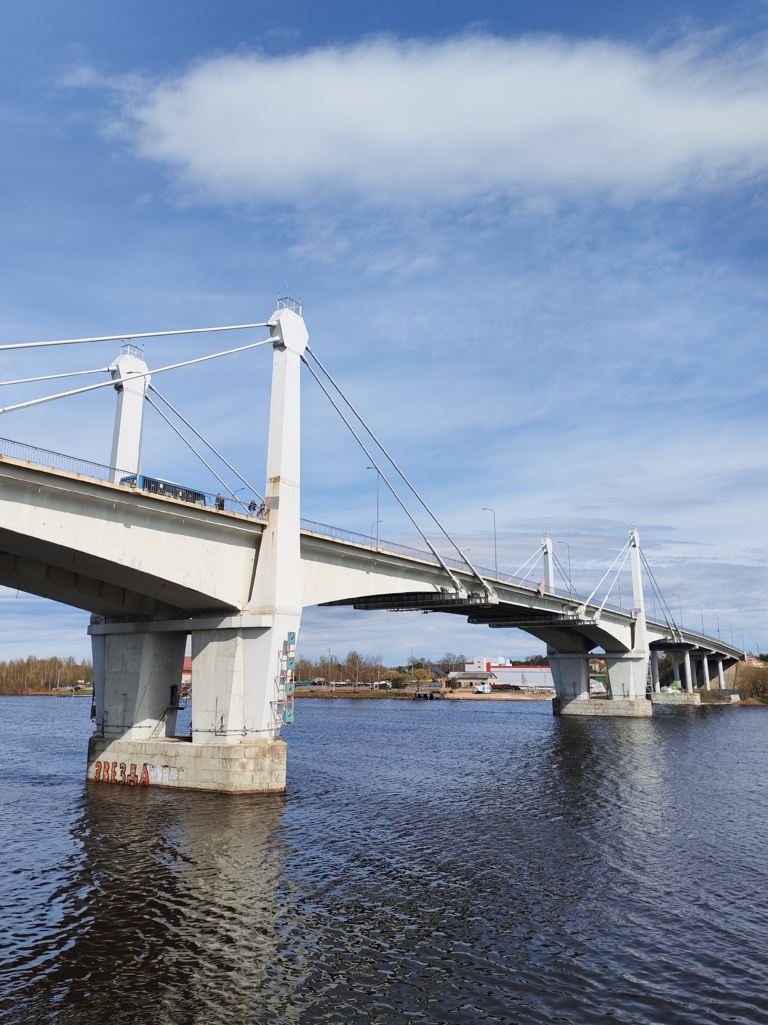 A daytime shot depicts a concrete bridge spanning a body of water, likely a river, under a sky with patchy clouds. The bridge's architecture features prominent concrete pillars and cable supports extending from the top of each pillar. There appear to be individuals and a vehicle on the bridge's surface. The river surface shows some wave activity, reflecting the light from the sky. On the riverbank, trees and structures, including some with red roofing, are visible. Graffiti is present on the base of one of the bridge's pillars. The city is Unknown, Unknown.