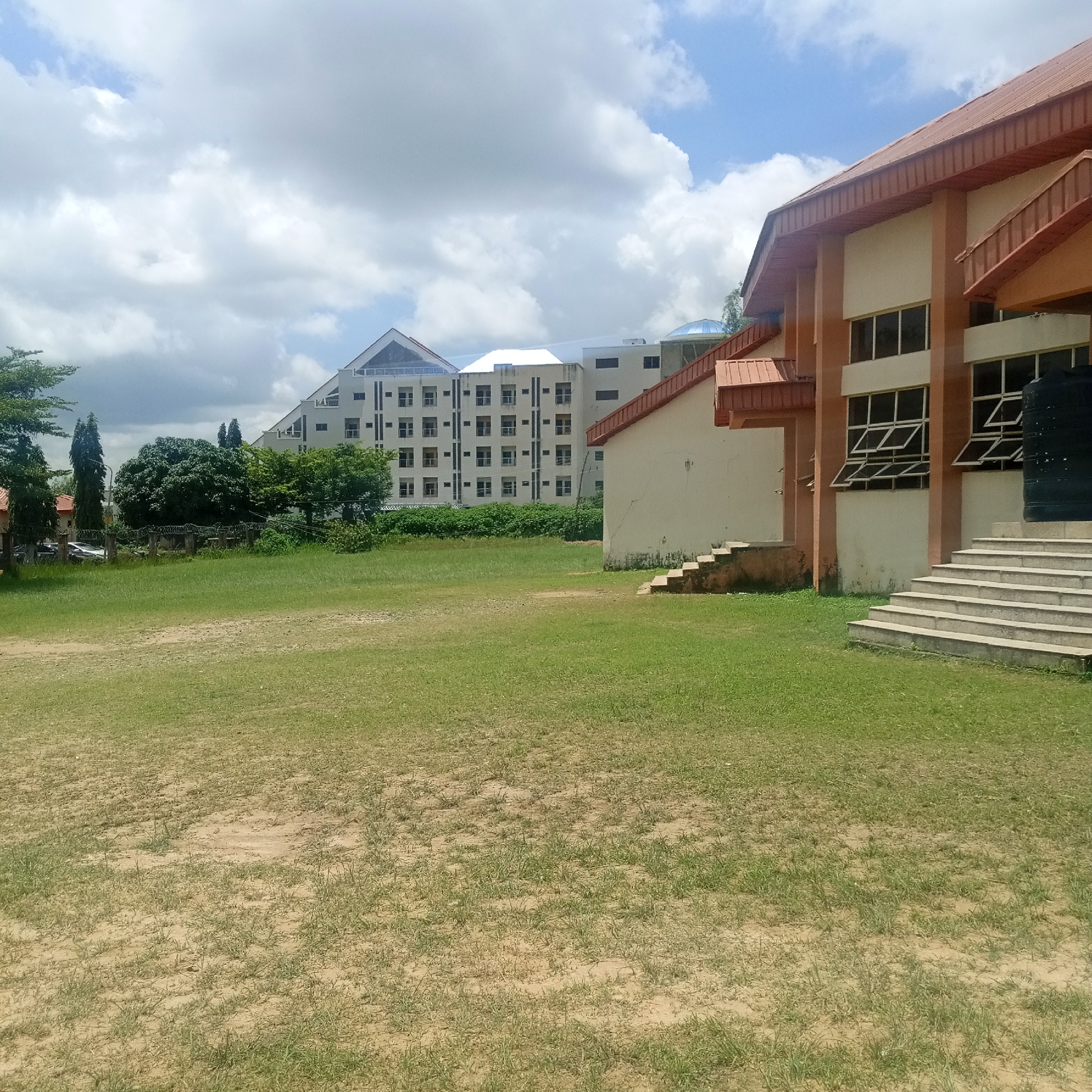 This is a photograph of the building complex of the National Centre for Petroleum Research and Development (NCPRD) located in Jos, Nigeria. The image captures the exterior of the facility on a partly cloudy day. In the foreground, there is a large, open grassy area with some patches of dry earth. To the right, a portion of a building with a reddish-brown roof and orange-colored exterior walls is visible. This building features a set of concrete stairs leading up to an entrance. Further in the background, a larger, multi-story building with a geometric, stepped facade and numerous windows dominates the scene. This building appears to be modern in design, with a white and light-colored exterior. Trees and vegetation are scattered throughout the image, particularly between the foreground and the background buildings. A barbed wire fence is also visible in the midground, separating the grassy area from the buildings. The sky is filled with white, fluffy clouds against a light blue background.