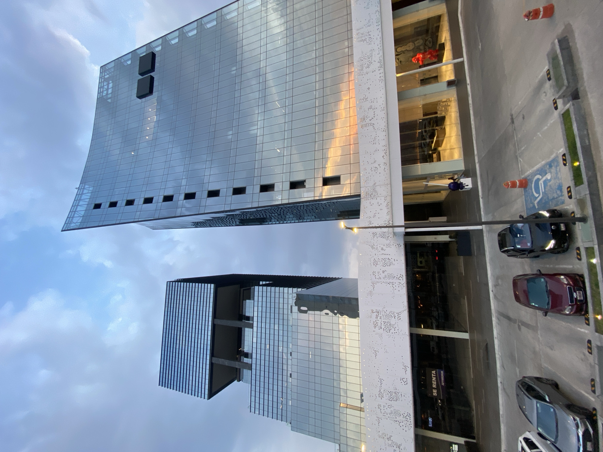 The image depicts an upward-looking perspective of modern architecture in Puebla, Mexico. Two glass-clad high-rise buildings dominate the scene. The taller building on the left has a reflective facade mirroring the sky, with notable dark square features near its top. The second building, appearing shorter and closer, also exhibits a glass facade. Beside it is a partially visible white, perforated vertical architectural element.
A street-level view is partially visible to the right, showing parked cars, including a red sedan and a black car in a designated handicap space. A sign with the words "SE RENTA" (FOR RENT) is reflected on the windows of the building on the right. Traffic cones are visible. The sky, partially obscured by the buildings, is a gradient of blue and cloudy.