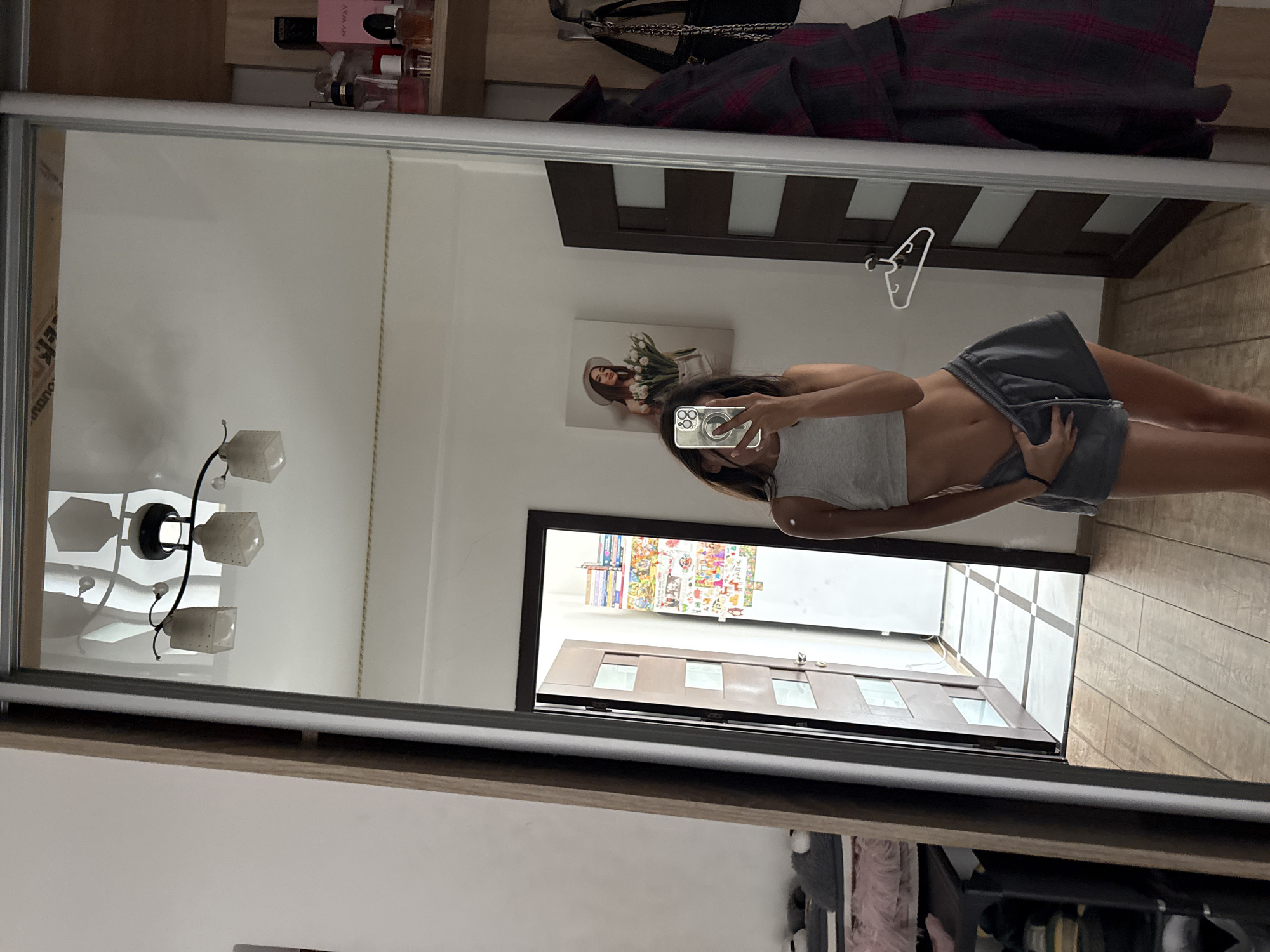 A full-length mirror reflection shows a young woman in a grey tank top and grey shorts. She is holding her phone in her right hand, using it to take the photo. Her left hand is resting on her shorts. The background of the reflection shows a room with a door, a painting on the wall, shelves with perfume bottles, and clothes hanging on a rack. The floor is tiled in a checkerboard pattern in front of the door and wooden planks elsewhere.