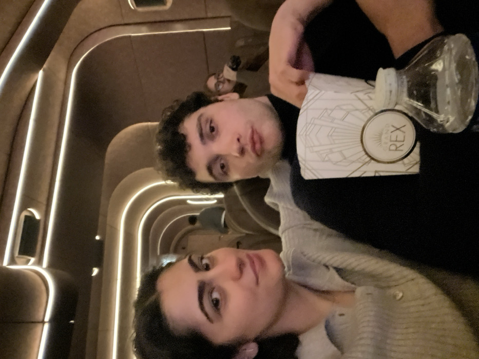 Two people pose for a selfie in what appears to be a movie theatre. The woman has dark hair and is wearing a gray cardigan. The man, with curly hair, is holding a container with "Grand Rex" printed on it, along with a water bottle. Another person can be seen blurred in the background. The theatre has a distinct design, featuring curved walls with integrated lighting.