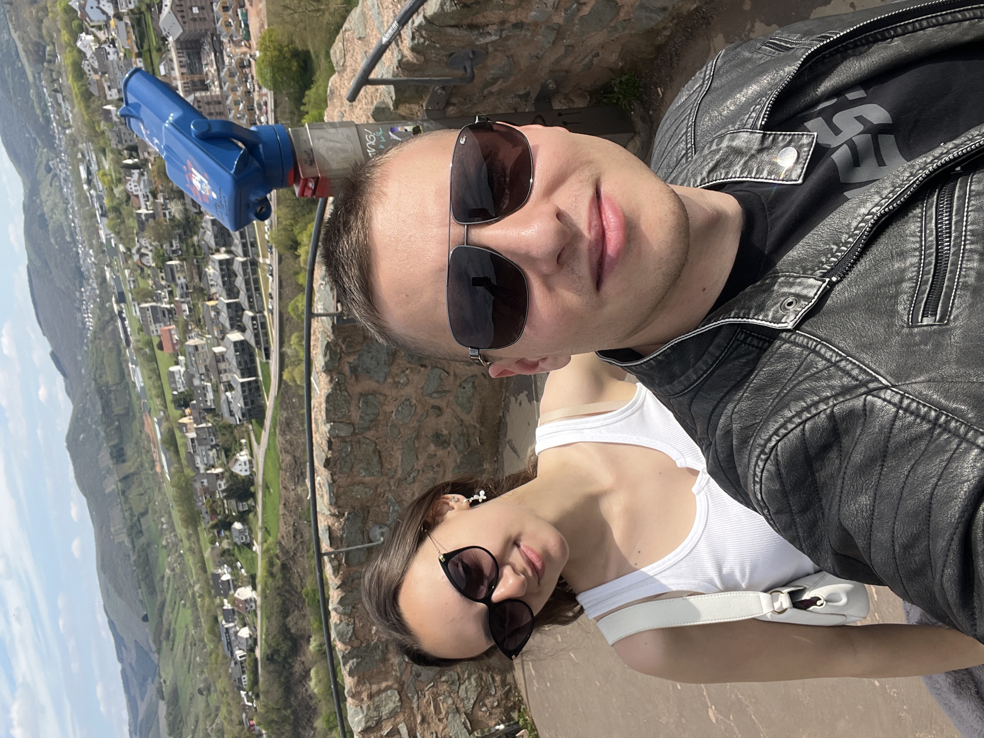 Two individuals pose for a selfie against the backdrop of Saarburg, Germany. A male figure, wearing a black leather jacket, is positioned in front, partially obscuring a woman wearing a white tank top and sunglasses. A blue coin-operated binoculars stands on a stone ledge, offering a view of the cityscape, which includes buildings nestled in a valley. The sky is bright. The setting appears to be a scenic overlook point. The stone ledge is made up of stone materials. The female is wearing an earring. She is wearing a white purse. The male is wearing a dark t-shirt underneath the jacket.