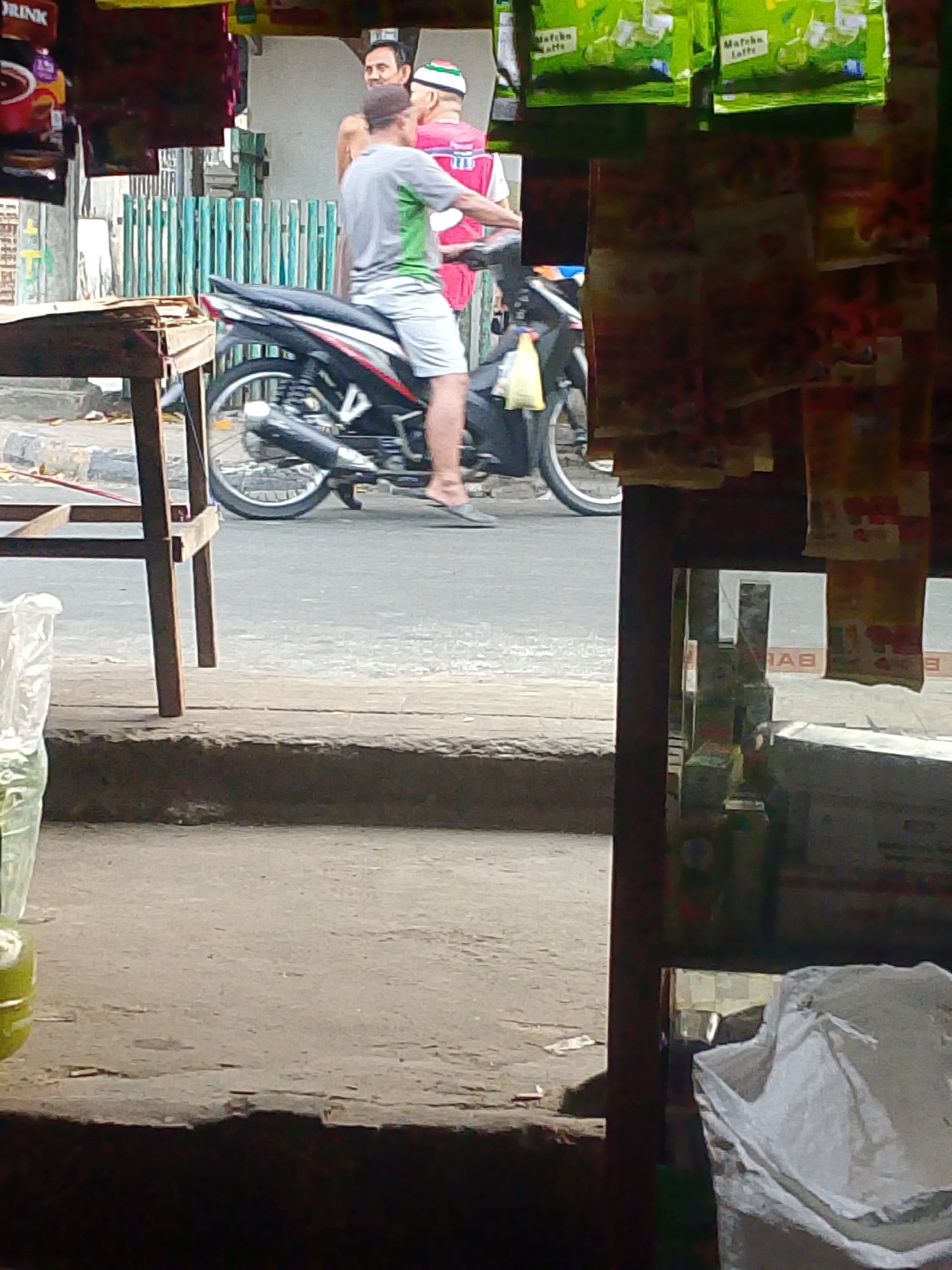 The image depicts a street scene in Palembang, Indonesia, likely near a market or shop. In the foreground, a wooden table and part of a storefront are visible. A motorcycle with a rider is central to the image. The rider, a man wearing a grey t-shirt with green accents and light-colored shorts, is seated on the motorcycle and appears to be looking to his right. Behind him, another man, partially visible, stands with his back to the camera, wearing a pink shirt and a green and white cap. The background shows a row of green vertical palings, suggesting a fence or barrier. Various items are displayed for sale, including packaged goods with visible Indonesian text and packaging. A large white bag is in the lower right of the frame, indicating a commercial setting where goods are stored or sold. The lighting suggests it is daytime.