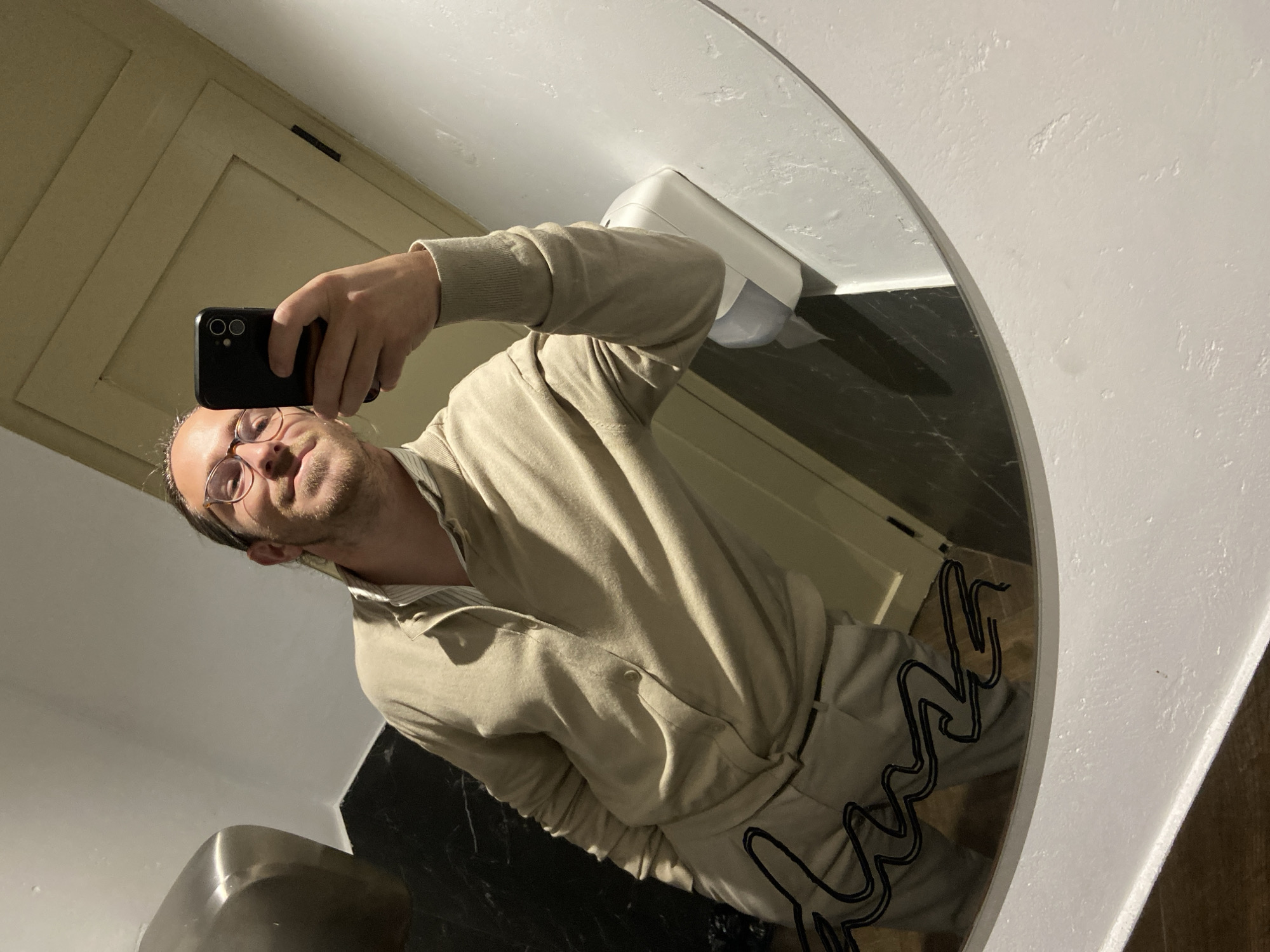 The image depicts a male subject taking a mirror selfie within a public restroom in Petrovac centar, Montenegro. The subject, a male appearing to be in his 20s to 40s, with light-colored hair pulled back and wearing glasses, smiles towards the camera reflection. He is dressed in a light beige cardigan over a light-striped collared shirt and light beige trousers, with his left hand in his pocket. He holds a black smartphone, featuring dual rear cameras, with his right hand to capture the image.
The environment includes a large, oval-shaped mirror with a white frame. Visible in the reflection are a metallic hand dryer on the upper left and a white wall-mounted dispenser (likely for soap or paper towels) on the right. The walls are predominantly white, with a section of dark, possibly black marble or granite tiling visible behind the subject's left side and reflected in the mirror. A light-colored wooden door with paneling is reflected in the upper right quadrant of the mirror. Black, wavy graffiti is present on the lower left surface of the mirror. The floor, partially visible in the mirror's reflection, appears to be dark-colored. Artificial lighting illuminates the scene.