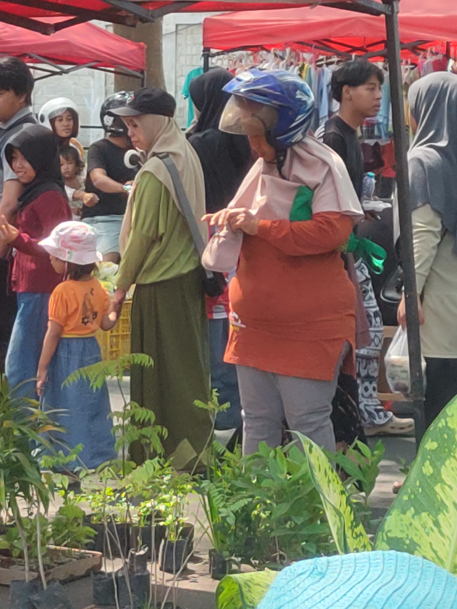 This is a medium shot photograph taken outdoors, likely at a market in Ngasem, Indonesia. The scene is crowded with people, many wearing headscarves or hijabs. In the foreground, a young child in a pink hat and orange shirt stands next to an adult. To the right, another person wears a blue helmet and an orange top. In the background, more individuals are visible, some appearing to be vendors with clothing displayed on racks under red awnings. The ground is paved, and there are plants in pots in the lower portion of the frame. The lighting suggests it is daytime.
