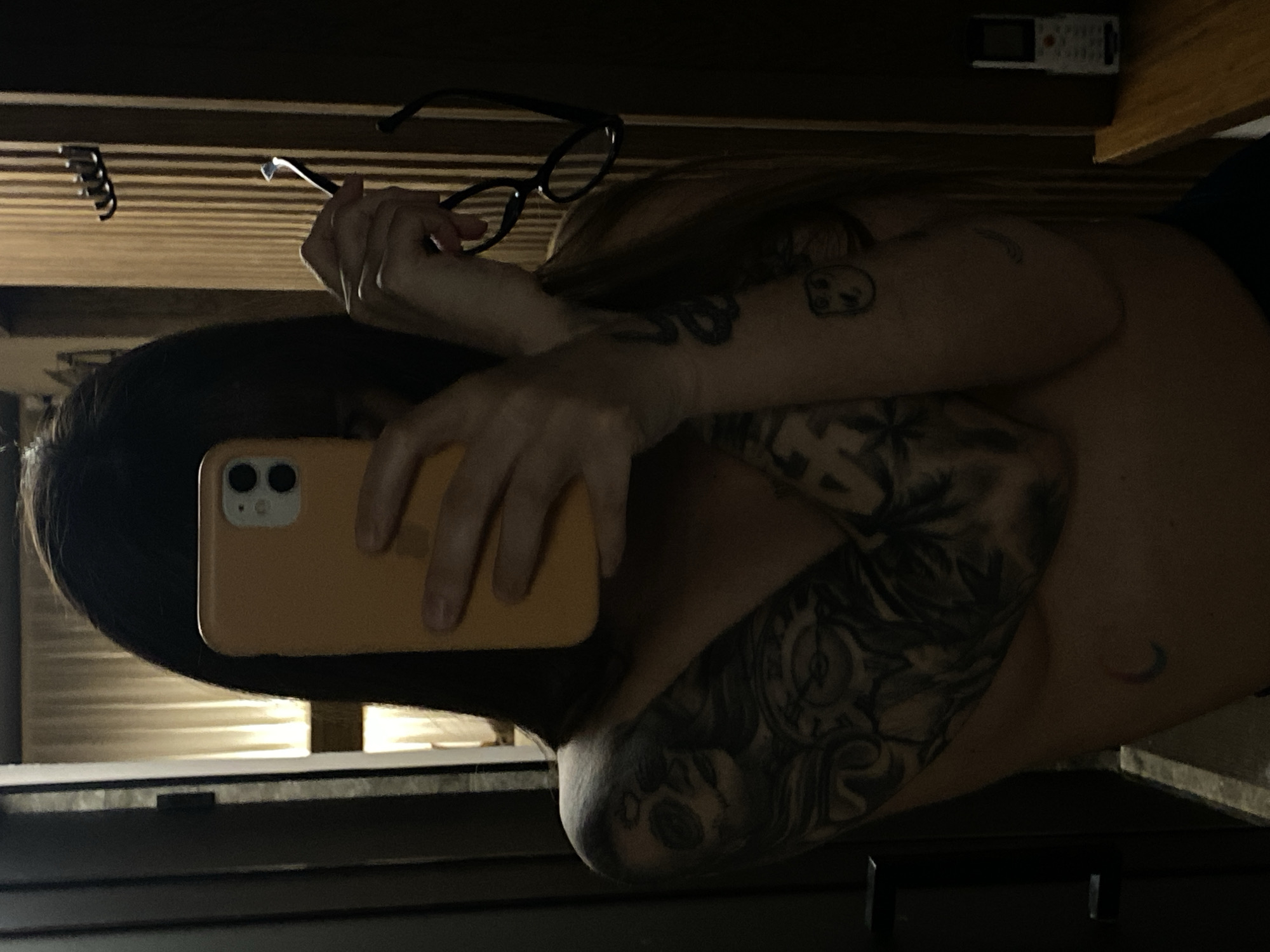 A person with long, dark hair is taking a selfie with an orange-colored smartphone. The person is holding a pair of black-rimmed glasses in their other hand. The person's arms are crossed, revealing tattoos on both arms. The environment is dimly lit and includes a window with blinds. A remote control is visible in the upper right corner. The location in the city of Votkinsk, Russia cannot be determined from the image.