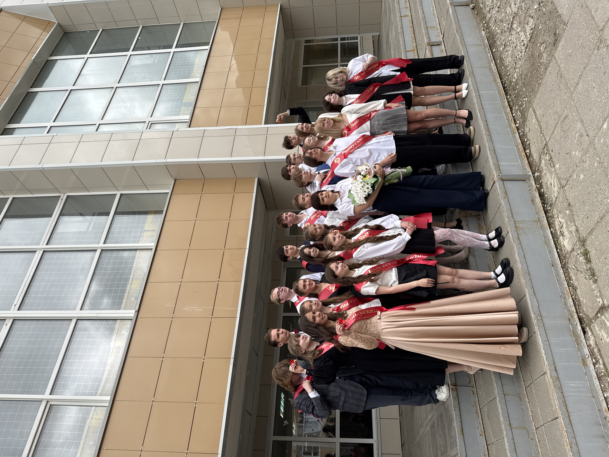 A group of approximately 20 graduating students is posing for a photo outside a building in Ivanovo, Russia. The students are standing on a set of outdoor stairs in front of a modern, multi-story building with large windows and beige paneling. Most of the students are wearing white shirts or blouses with dark skirts or pants, and they are all wearing red sashes with white Russian text, identifying them as graduates. One student is holding a bouquet of white flowers. The scene appears to be a celebratory occasion, likely a graduation ceremony or photo shoot. The exterior suggests an educational institution, possibly a school or university. The day is overcast, casting a soft light on the scene. The building's architectural style appears modern and functional. The students are oriented in a line, facing upwards, with their faces visible. The surrounding pavement is visible, suggesting an outdoor setting.