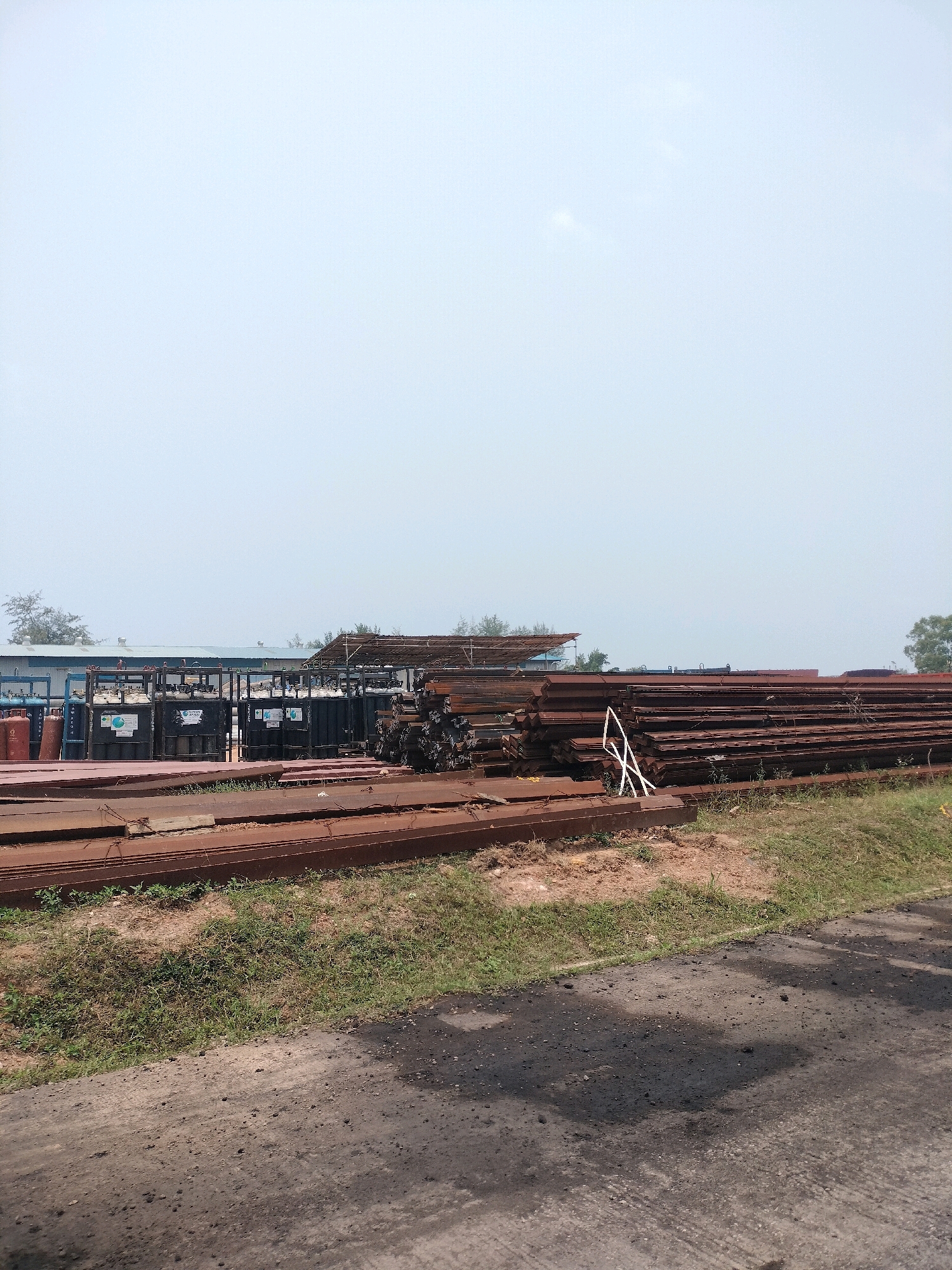 This image depicts an outdoor storage area for industrial materials, possibly in a port or manufacturing district in Batam, Indonesia. The foreground shows a rough, unpaved or asphalt surface with some debris. In the middle ground, large stacks of rusty metal beams and plates are piled high. Behind these metal stocks, there are several large, dark metal cages or racks containing what appear to be gas cylinders, some red and others black, with labeling visible on some. Further in the background, industrial buildings with corrugated metal roofs are visible, surrounded by sparse vegetation and trees, under a hazy, pale sky. The scene suggests a commercial or industrial site where materials are stored before or after processing, shipping, or use. There are no people visible in the image.