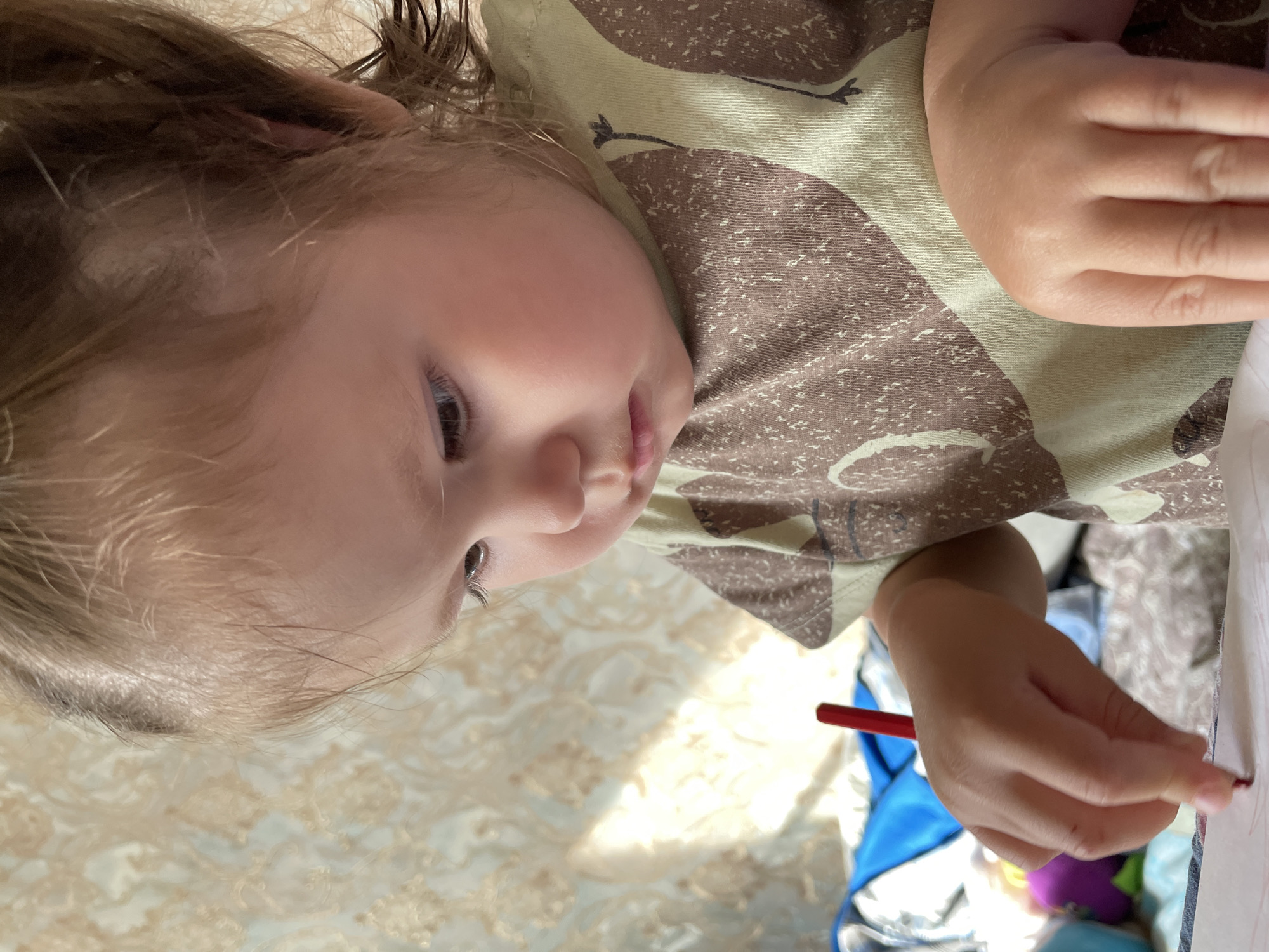The media file depicts a young child, likely a toddler, engaged in drawing or coloring indoors in Izhevsk, Russia. The scene shows the child's upper body from an overhead perspective, with their face turned downwards in concentration. The child has light brown or blonde hair and wears a light green t-shirt with a brown, abstract patterned design.
In the child's right hand, a red crayon or colored pencil is held, actively marking on a piece of paper or in a coloring book, which is partially visible. The background consists of a light-colored, intricately patterned fabric, possibly a bedspread or blanket, suggesting a home setting. A distinct patch of bright sunlight illuminates part of the background, indicating it is daytime. Blurred objects, including hints of blue and purple, are visible beneath the child's hand, possibly toys or other belongings. The child's expression conveys focus and engagement. No text is visible in the image.