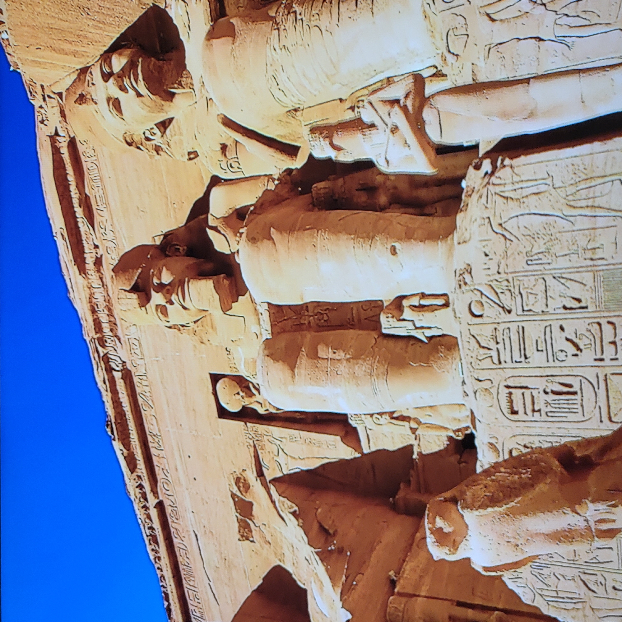 The image depicts a section of an ancient Egyptian monument, possibly a temple or tomb facade. The structure is constructed of light brown stone, with detailed carvings of human figures and hieroglyphics. The figures appear to be seated and are carved in relief. The hieroglyphics are inscribed on the surrounding surfaces. The background is a clear, bright blue sky. There is no evidence in the image to suggest that it is located in Yerevan, Armenia.