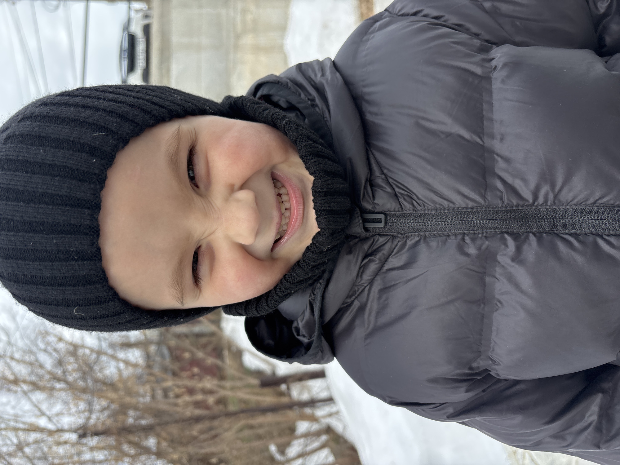 A child wearing a black knitted hat and black puffy winter jacket is smiling at the camera. The child is outside in a winter scene with snow visible in the background. Trees, partially obscured by snow, are also visible. The location is speculated to be in the city of Chaikovsky, Russia. The child's head is slightly tilted, and their teeth are visible in their smile.