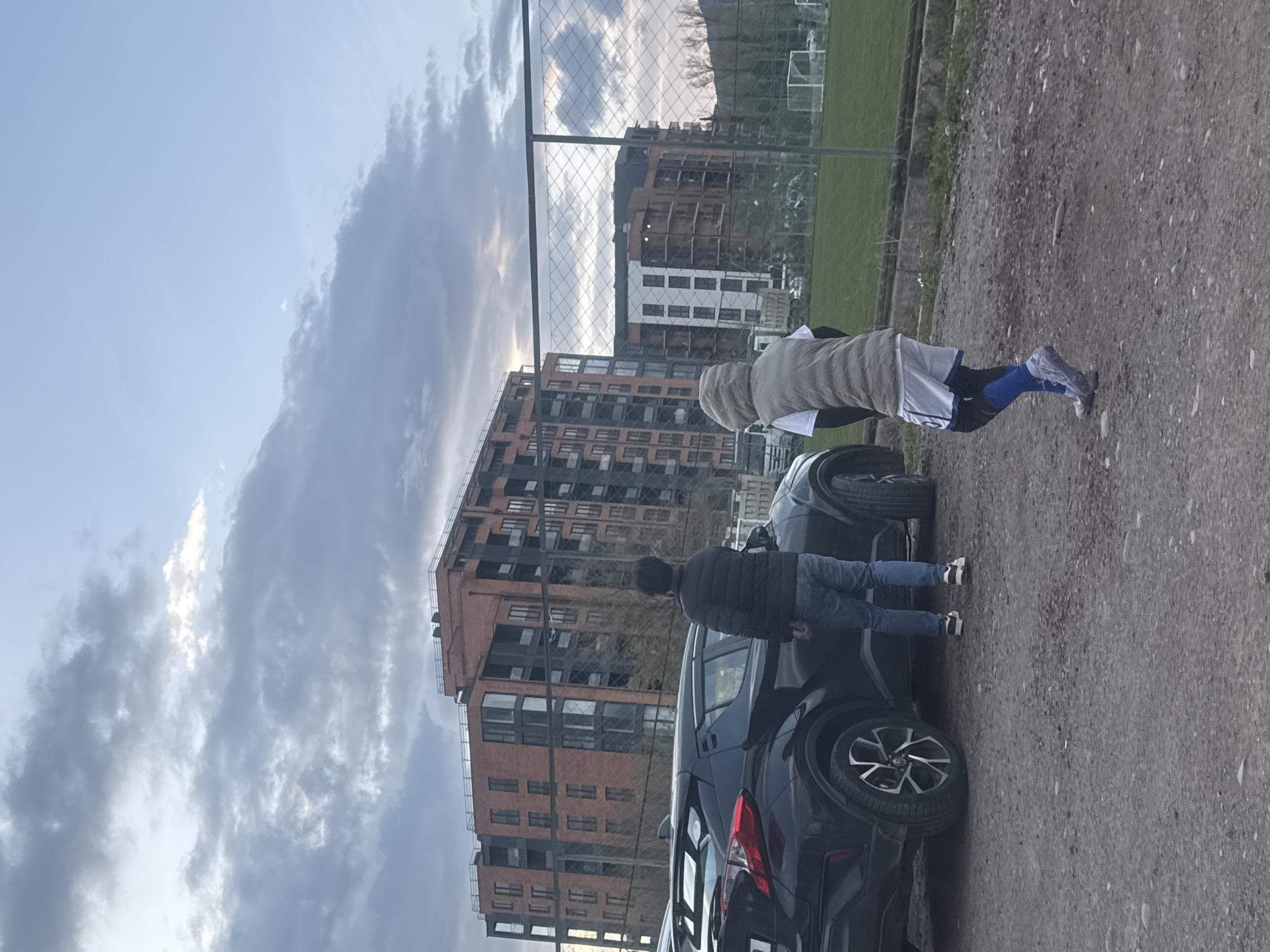 A horizontal image shows two people standing next to parked cars near buildings and a field. The sky above is filled with clouds. One person is wearing a dark jacket and jeans. The other person is wearing shorts, a vest, and compression sleeves on their legs. The cars are parked on a gravel surface. A chain-link fence separates the parking area from a grassy field in the background, where a soccer goal is visible. Buildings are behind the field. The scene appears to be in an urban area, possibly in Tbilisi, Georgia, given the architecture and general setting.