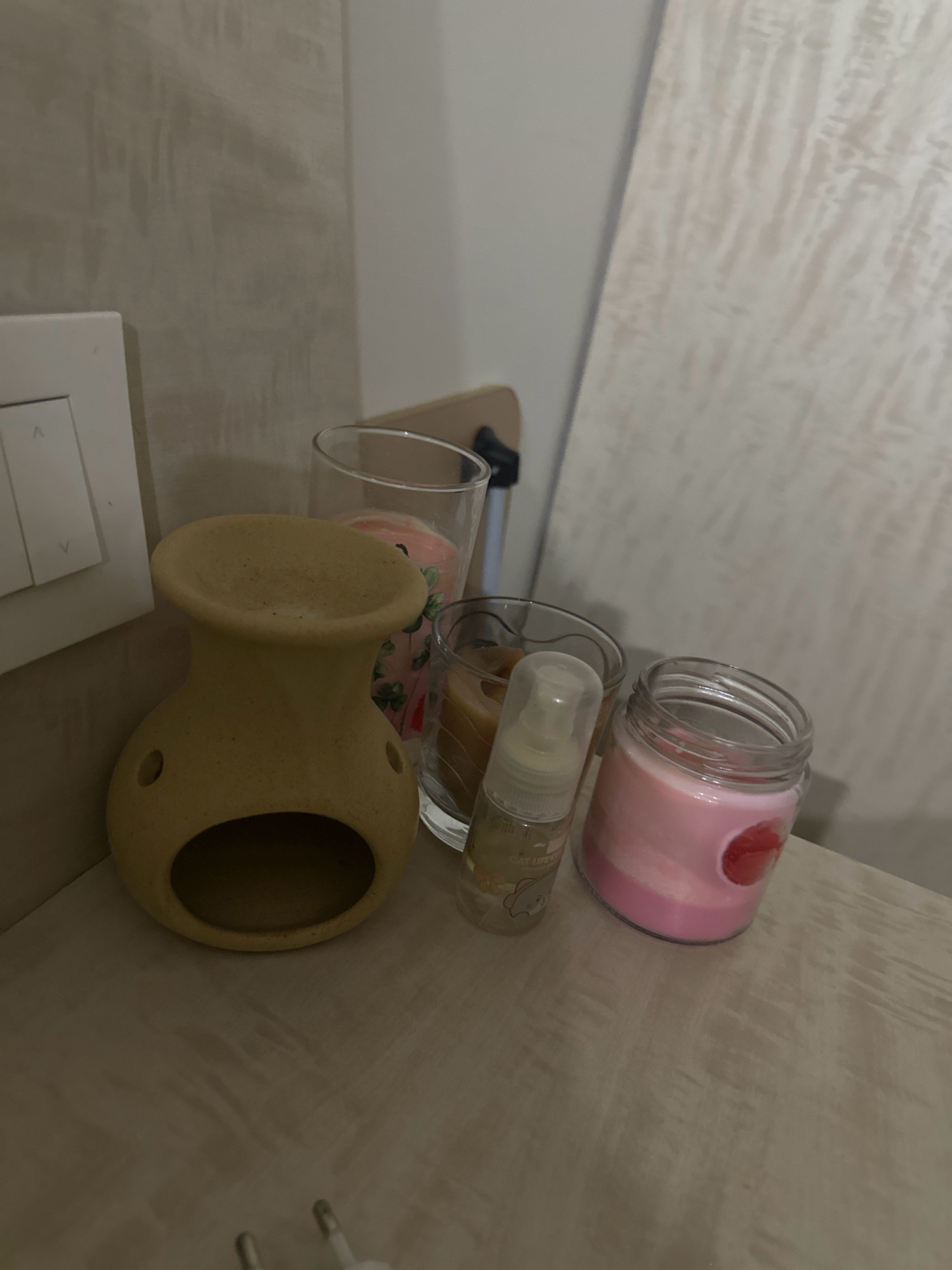 In Lalitpur, Nepal, on a wooden surface, several decorative items are arranged. In the foreground, a beige ceramic oil diffuser with a wide opening at the top and two circular holes on the sides sits to the left. Next to it, a small, clear spray bottle with a white cap contains a yellowish liquid and has a cartoon drawing of a cat on its label. To the right of the spray bottle, a pink candle in a glass jar is visible, with a red circular design embedded in the wax. Behind these items, two clear glasses contain candles. One glass holds a candle with what appears to be a pink floral pattern and small green clovers. The other glass holds a candle with a reddish-brown hue. A white light switch plate is mounted on a textured gray wall in the background, and a light-colored, vertically oriented object, possibly a piece of wood or paneling, is leaned against the wall to the right. The lighting is dim, creating shadows and highlighting the textures of the surfaces.