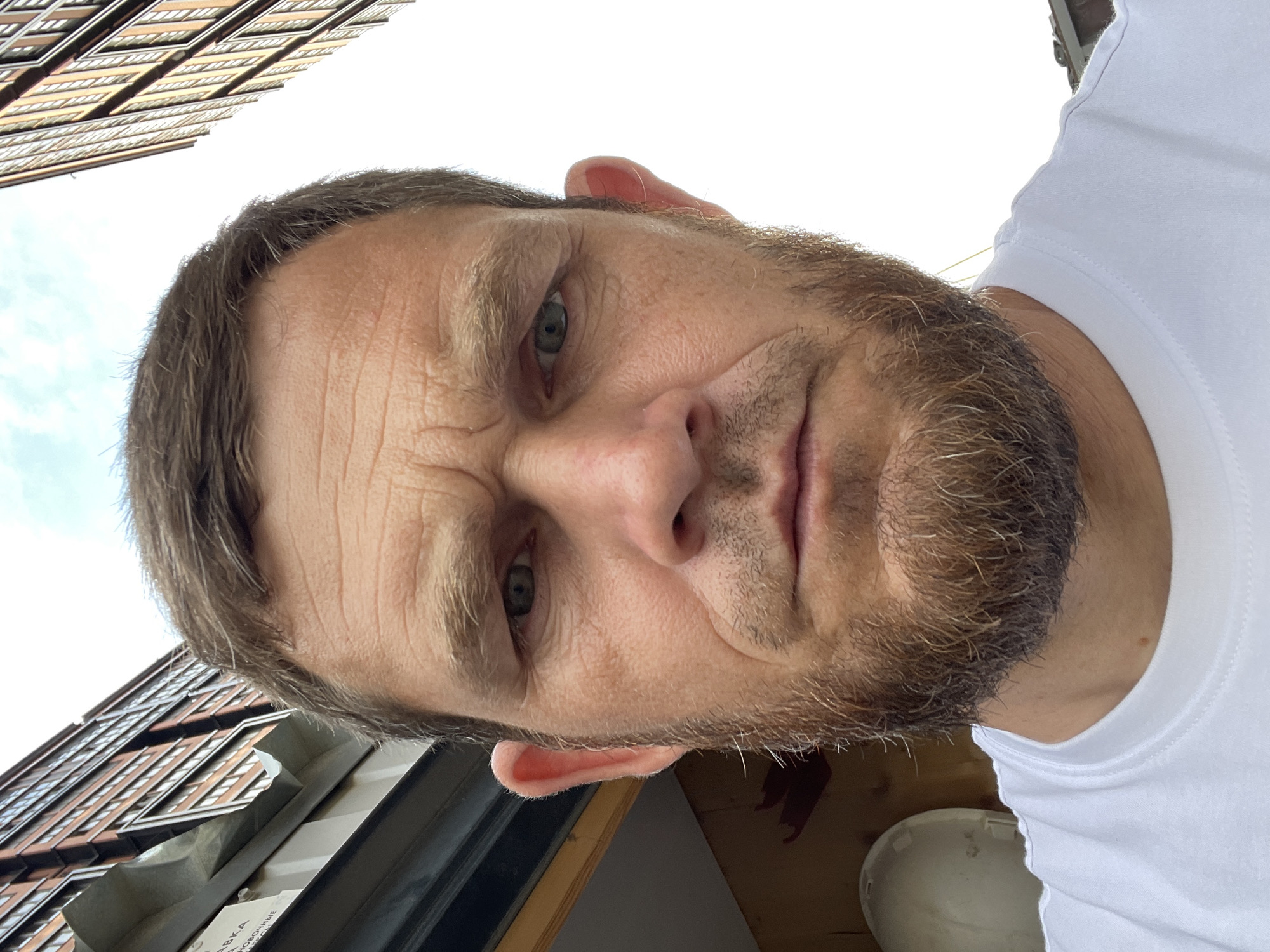 A man with short brown hair and a beard is shown in a close-up, outdoor, head-and-shoulders shot. He is wearing a white t-shirt. The background consists of a light sky and two tall buildings. One building, on the left, is made of red brick with many windows and is angled towards the camera. The other building, on the right, is also tall and appears to have a similar facade. A portion of a white object, possibly a hard hat, is visible to the left of the man's face. The man's expression is neutral. The image was taken in the city of Krasnodar, Russia.